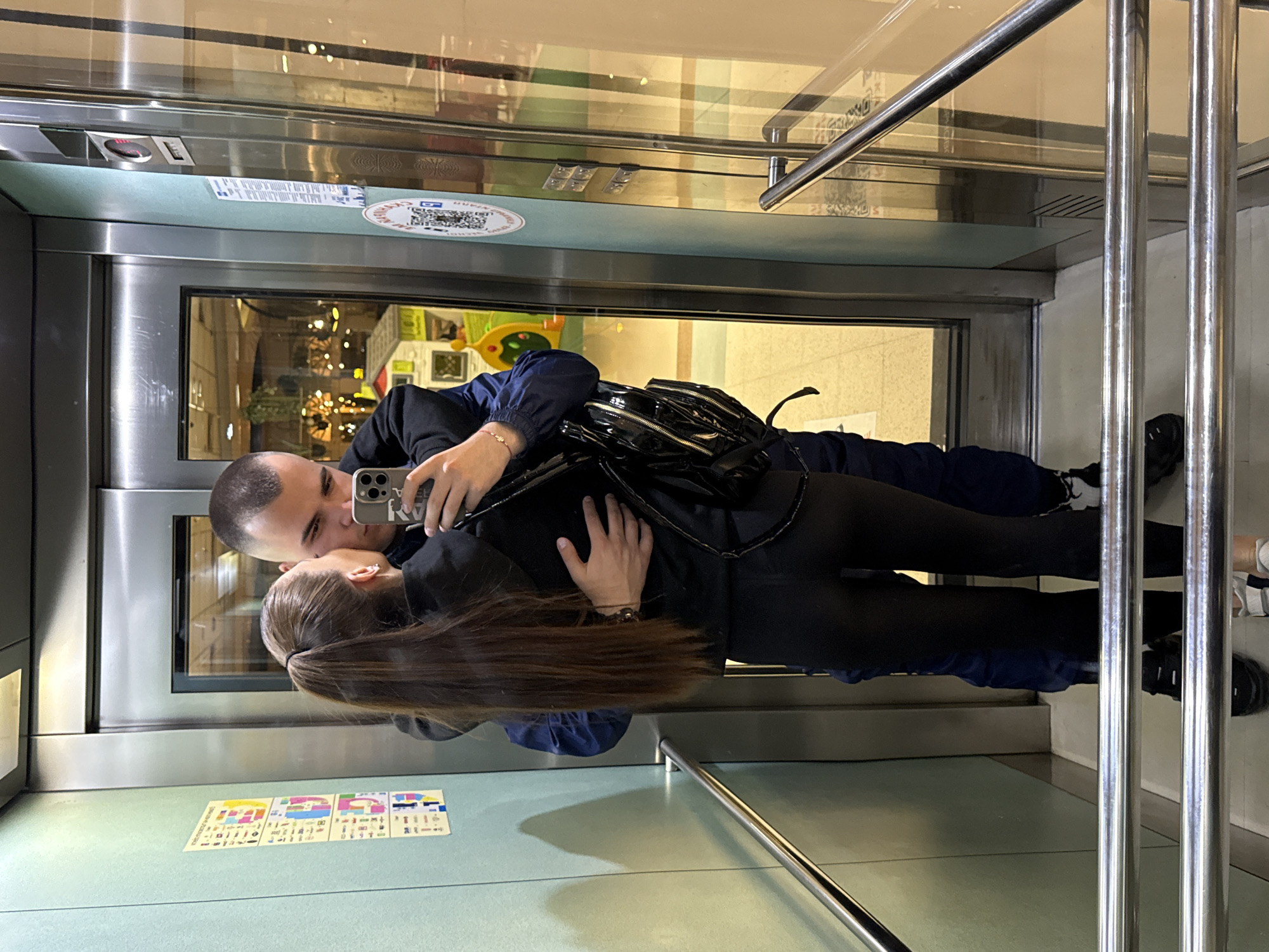 A man and woman are taking a selfie in an elevator. The man is holding a smartphone and is looking at the screen. The woman is looking at the man and is holding his arm. They appear to be posing for the photograph. The elevator has a mirrored wall, stainless steel bars, and information sticker/placard adhered to the inside wall. Outside the elevator appears to be a public space possibly a shopping mall.