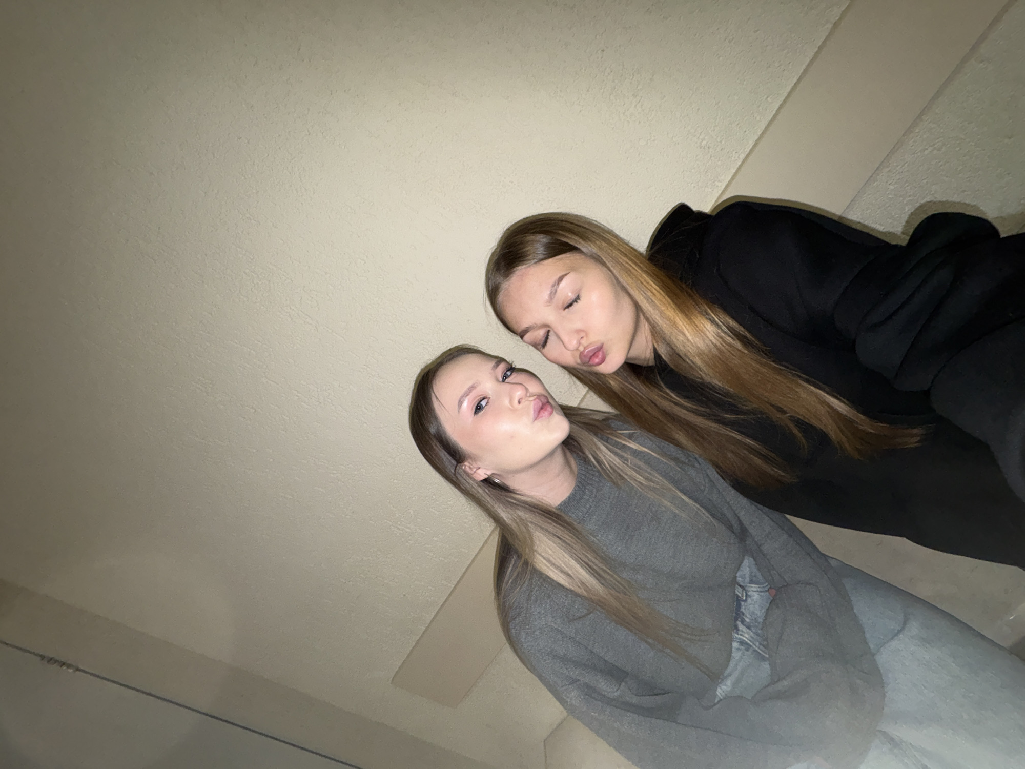 Two young women are posing for a photo. The woman in the foreground is wearing a grey sweater and light-wash jeans, and the woman in the background is wearing a black hoodie. Both women have long, light-colored hair and are making a kissing face. The photo appears to have been taken indoors, against a wall with what appears to be a white trim. The photo's location is Chaikovsky, Russia.