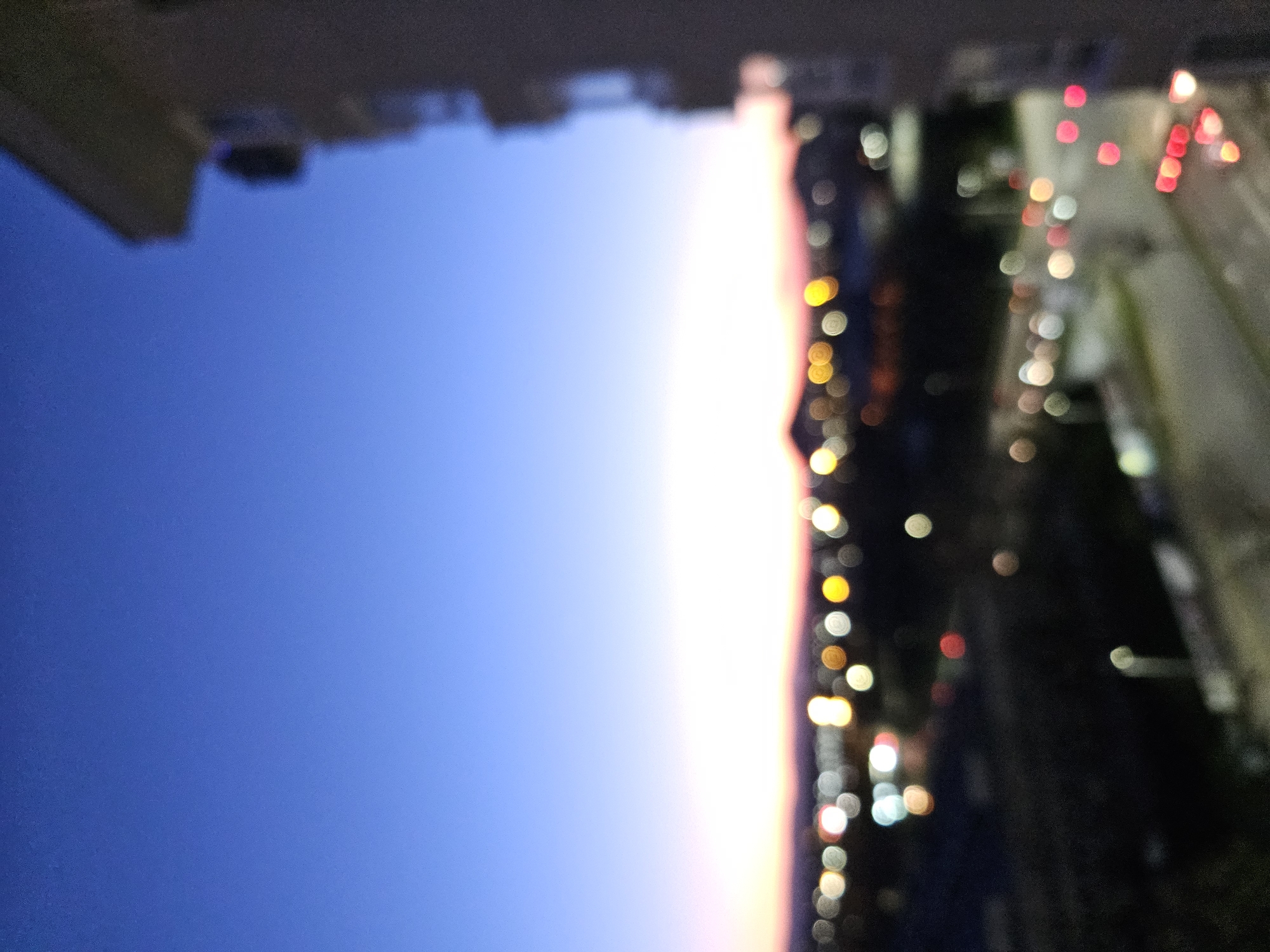 The image shows an out-of-focus cityscape at dusk, likely taken from a high vantage point. The left side features a blue sky transitioning to a faint pink horizon line. The right side displays blurred lights, suggestive of city streets and buildings at night. Red and white bokeh indicate tail lights of vehicles, and yellow bokeh represents streetlights or building lights. A portion of a building is visible at the top of the image. Without additional visual context, it's not possible to confirm the precise location in Plovdiv, Bulgaria.