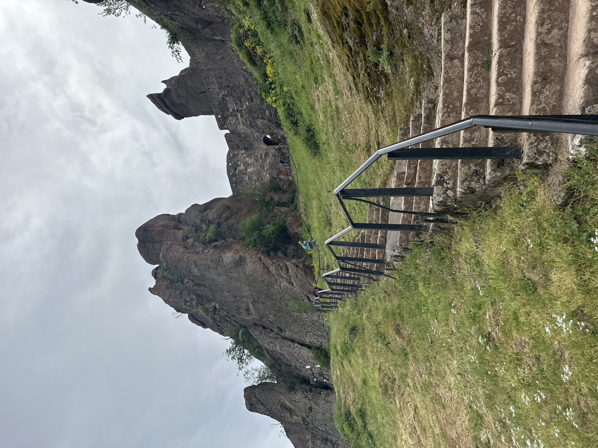 The image depicts a portion of the Belogradchik Fortress, a historic fortification located in the city of Belogradchik, Bulgaria. The fortress is situated amidst unique rock formations.
The scene shows a set of stone steps, equipped with a black metal handrail, leading upwards. The steps are adjacent to a steep, grassy slope. The rock formations, typical of the Belogradchik Rocks, are visible on the left side of the frame, extending towards the sky.
Two people are visible on the stairs, presumably tourists exploring the fortress. The sky is overcast, suggesting cloudy weather conditions. The overall environment is green, with grass and vegetation covering the slopes surrounding the rock formations and the fortress walls.