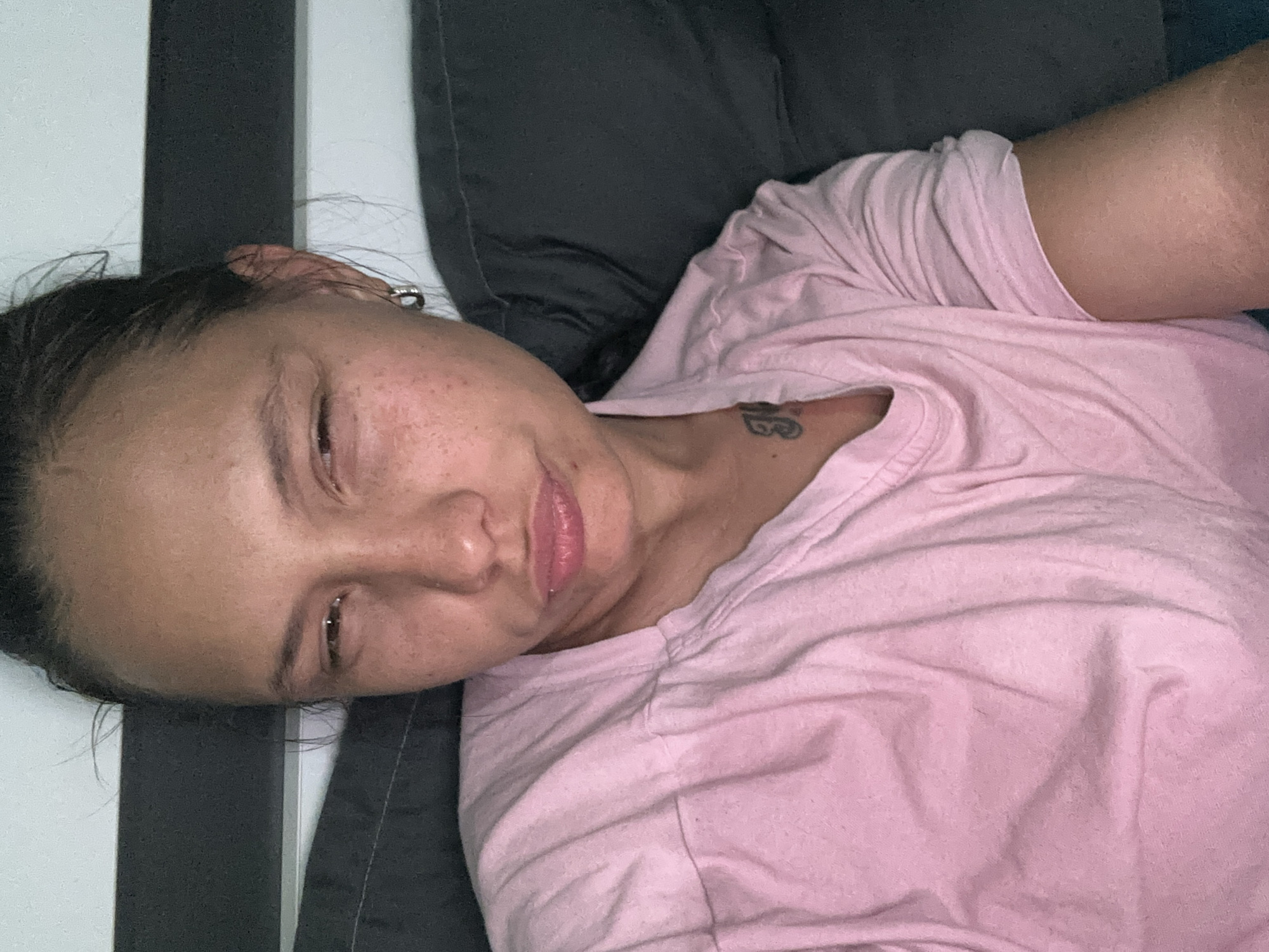 The image displays an adult female in a close-up, chest-up view within an indoor setting. Her dark hair is pulled back, with a few strands loose around her face. She has medium skin tone with visible freckles or minor blemishes on her cheeks and nose. Her eyes are squinted, and her lips are pursed in a pout, conveying an expression of mild annoyance, skepticism, or discomfort. A small silver-colored hoop earring is visible in her right ear. A dark tattoo, possibly text or a symbol, is partially visible on her upper chest, near her collarbone, beneath the neckline of her shirt. She is wearing a light pink, short-sleeved t-shirt, which appears to be made of cotton and exhibits some wrinkles. The background consists of dark grey or black fabric, likely upholstery from a bed or couch, directly behind and to the sides of her. Above this dark fabric, on the left, a light-colored wall or partition is visible with a thin dark horizontal stripe. The lighting is diffused and even. The scene's specific location within Sofia, Bulgaria, is not discernible from the visual information provided in the image itself.