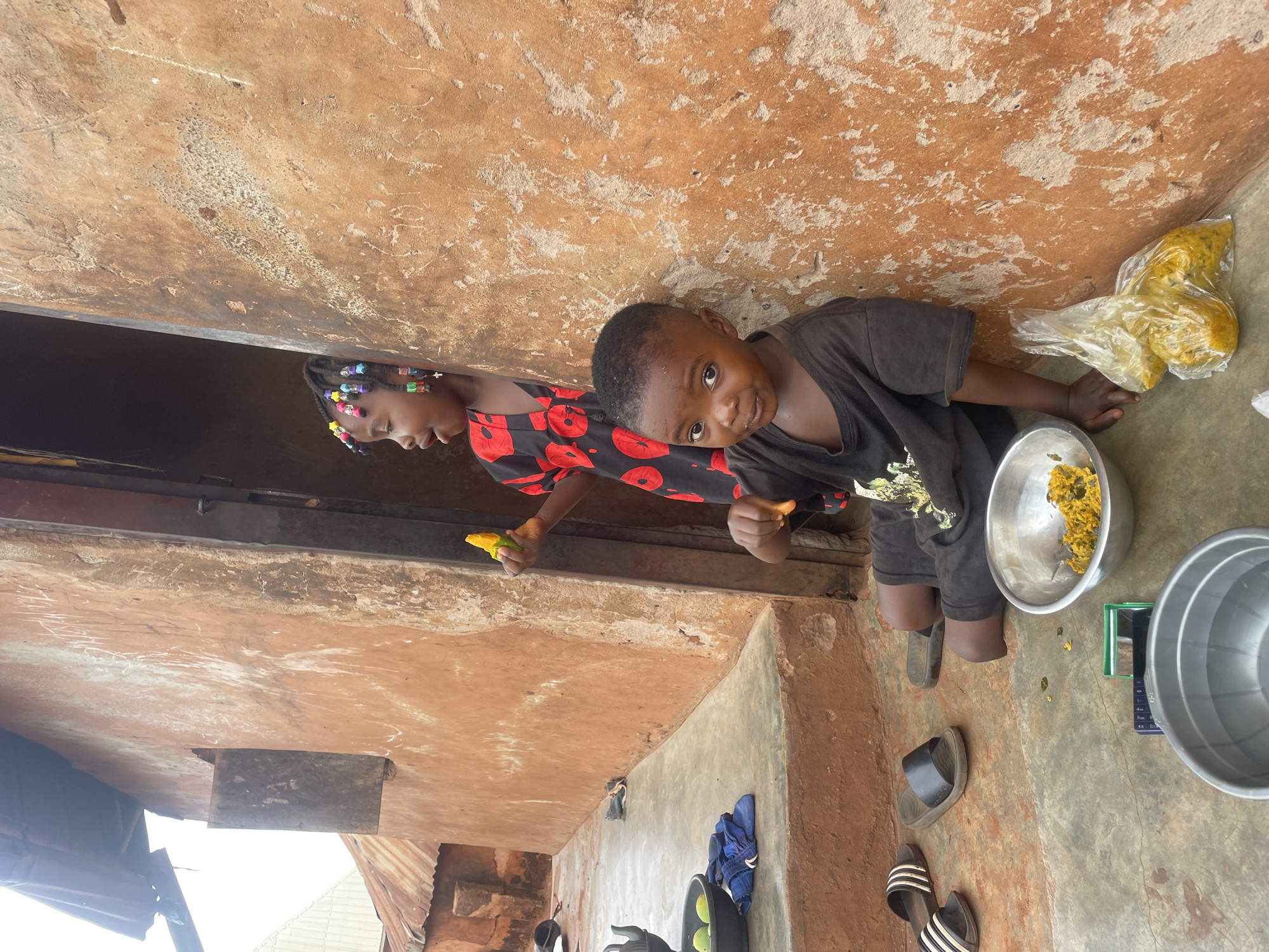 In Ogbe, Nigeria, two children are outside near a building with textured orange walls. A boy in a black t-shirt looks at the camera while holding a piece of food. Beside him is a silver bowl filled with food. A girl with colorful beads in her hair, wearing a red and black dress, holds food in her hand. Nearby are other silver bowls, a plastic bag containing food, sandals, and blue clothes. The building has a tin roof.