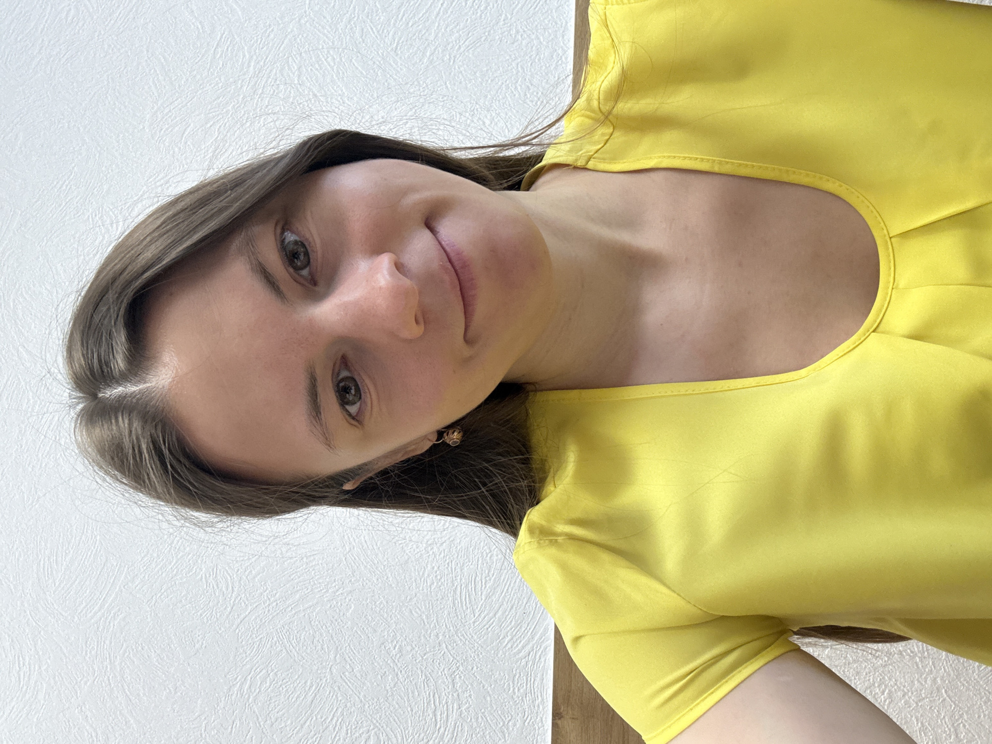 A person, likely female, is shown in a close-up shot against a white wall. The individual has light brown hair and is wearing a yellow top with a round neckline. The photo was taken indoors, and the subject appears to be looking directly at the camera with a slight smile. Specific details placing this image in Chaikovsky, Russia, are not discernible from the image itself.