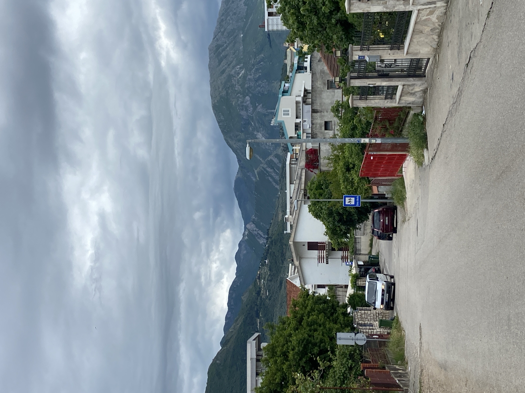 The image captures a street scene in Bar, Montenegro. The view is along a paved road lined with buildings of varying styles and sizes. The architecture is a mix of residential structures, most appearing to be multi-story houses with light-colored facades and some with red-tiled roofs. Green trees and shrubs are scattered throughout, softening the urban environment.
A prominent mountain range dominates the background under a cloudy sky. The clouds are dense and layered, suggesting overcast conditions.
Several vehicles are present on the road. A burgundy vehicle and a white vehicle are visible. Street signs with lettering, partially obscured by foliage, are placed along the roadside. One sign has a distinctive white symbol on a blue background, and is in a protected location.