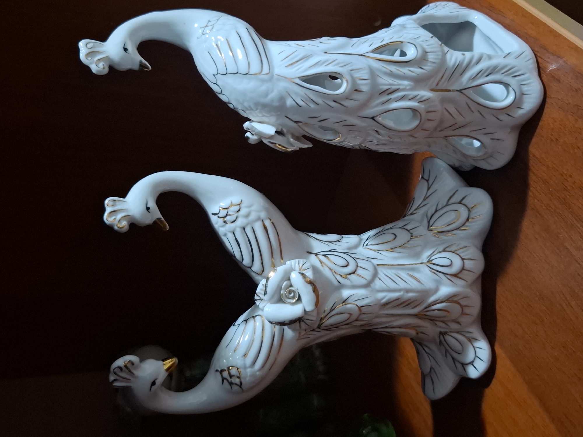 Two white porcelain peacock figurines with gold accents are displayed on a wooden shelf. The figurine on the left is a pair of peacocks facing each other, connected at the base and adorned with a white rose with gold detailing. The figurine on the right is a single peacock with an open tail, also decorated with gold lines resembling feathers. The background is dark and appears to be the interior of a room. The lighting suggests an indoor setting with a light source from the front. The scene is static and there are no people or animals present. There is no information available regarding the city of Almaty, Kazakhstan.