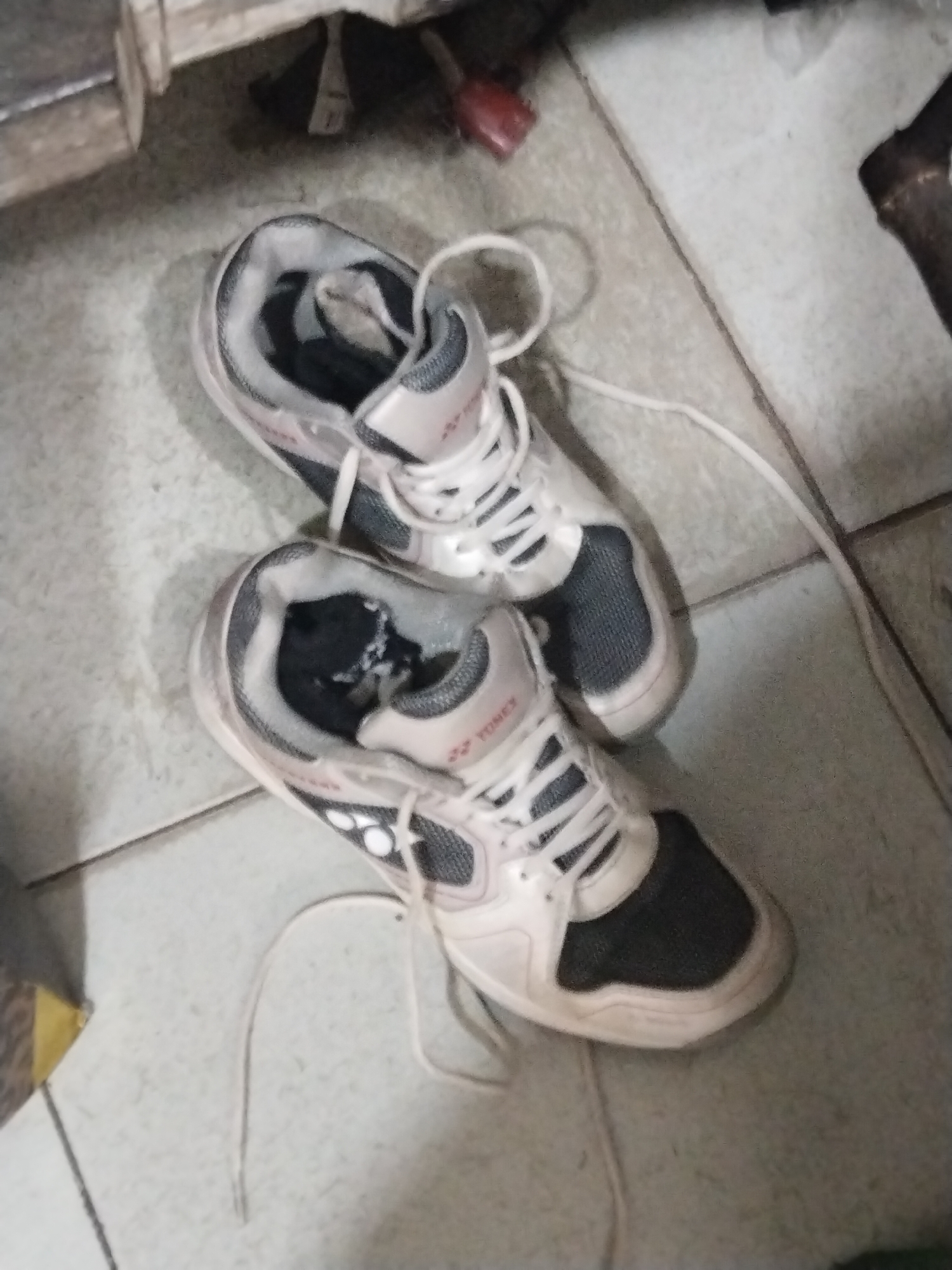 Two white and grey Yonex athletic shoes are on a tiled floor. The shoes are side by side, with the laces untied and spread out. The right shoe is slightly above and to the right of the left shoe. The floor tiles are a light grey color. In the background, there is a wooden object to the left and a dark, cylindrical object to the right. The scene appears to be indoors, possibly in a residential setting.
