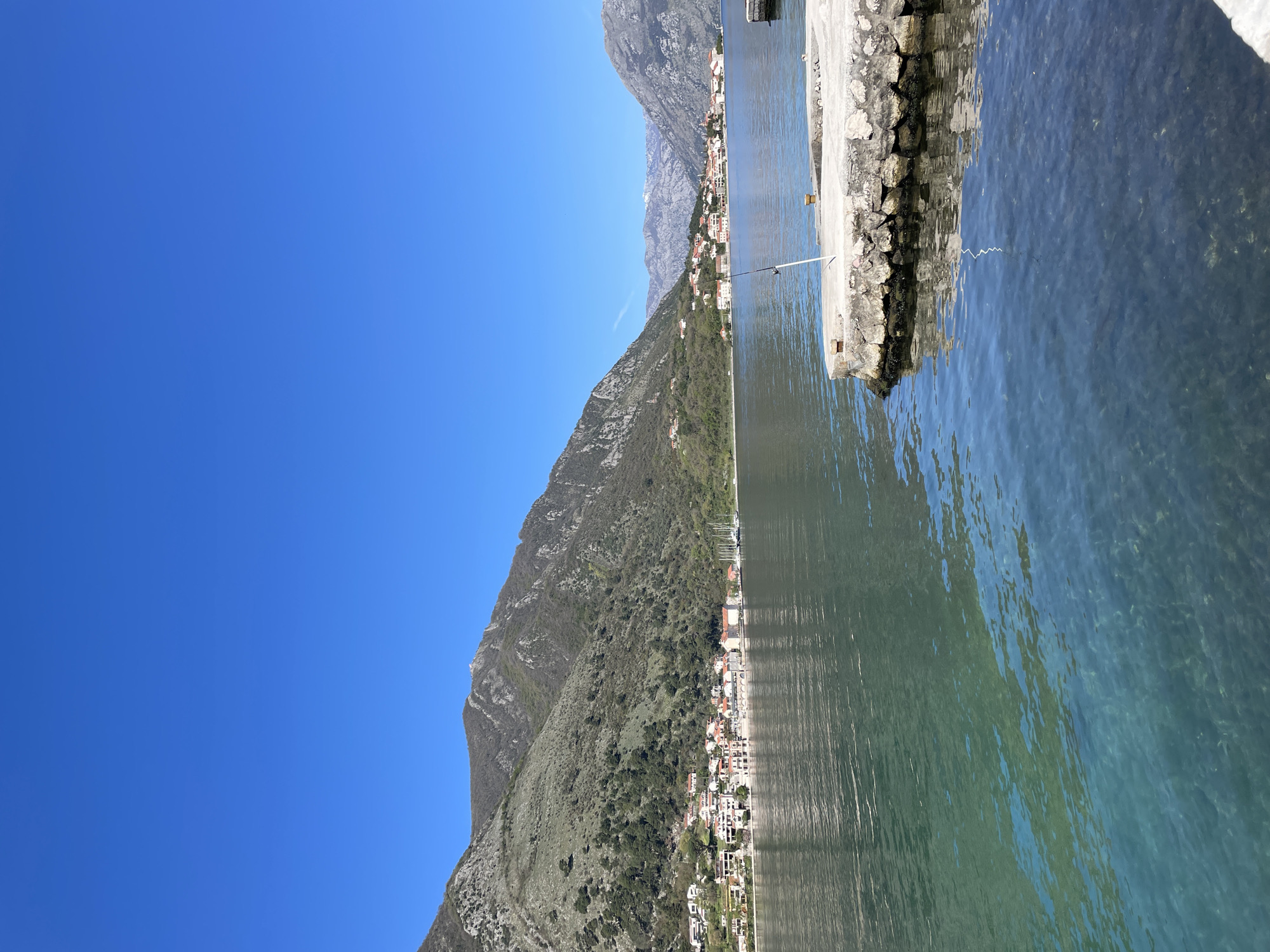 The image captures a view of Dobrota, Montenegro, featuring a waterfront perspective. Clear turquoise water fills the foreground, meeting a stone pier on the right. The town stretches along the coastline at the base of a mountain. Buildings with red-tiled roofs are visible among green vegetation. The sky is a clear, bright blue.