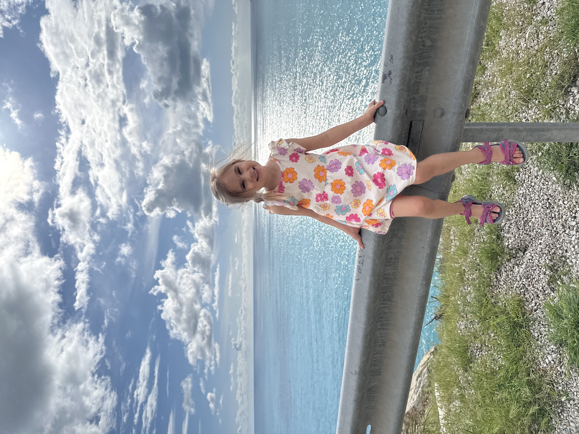 A young girl with blonde hair in pigtails stands on a gray guardrail overlooking a body of water, presumed to be the Mediterranean Sea, which has a sparkling sun reflection. The sky is blue with cumulus clouds. She is wearing a light-colored dress with floral patterns and sandals. The guardrail separates her from the water below. Beyond the guardrail is grass and small stones. The location is likely near or in Fethiye, Turkey, given the prompt.