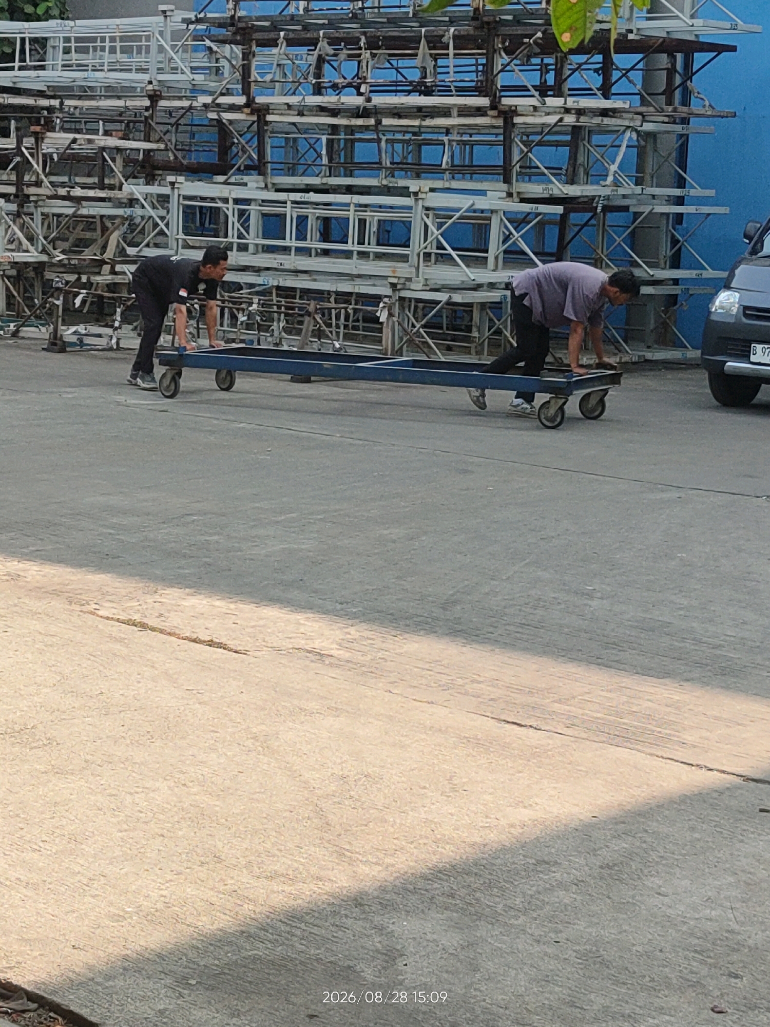 Two men are moving a long, blue, metal frame on wheels. The frame has two sets of wheels, one at each end. The man on the left is wearing a black t-shirt and black pants, and the man on the right is wearing a grey t-shirt and black pants. They are both pushing the frame. In the background, there are stacks of metal scaffolding and a blue wall. A dark colored van is parked to the right of the frame. The image appears to have been taken outdoors on a sunny day. The timestamp at the bottom right of the image reads "2026/08/28 15:09".