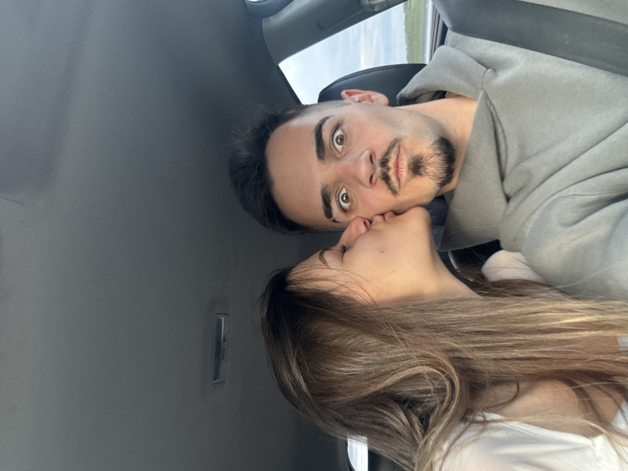 Two people are in a car. A woman with long brown hair is kissing a man. The man has dark hair, a beard, and is wearing a gray hoodie. They are inside a car with a gray interior. The location is unknown. The image does not provide sufficient information to determine if the scene is in the city of Chokoba, Bulgaria.