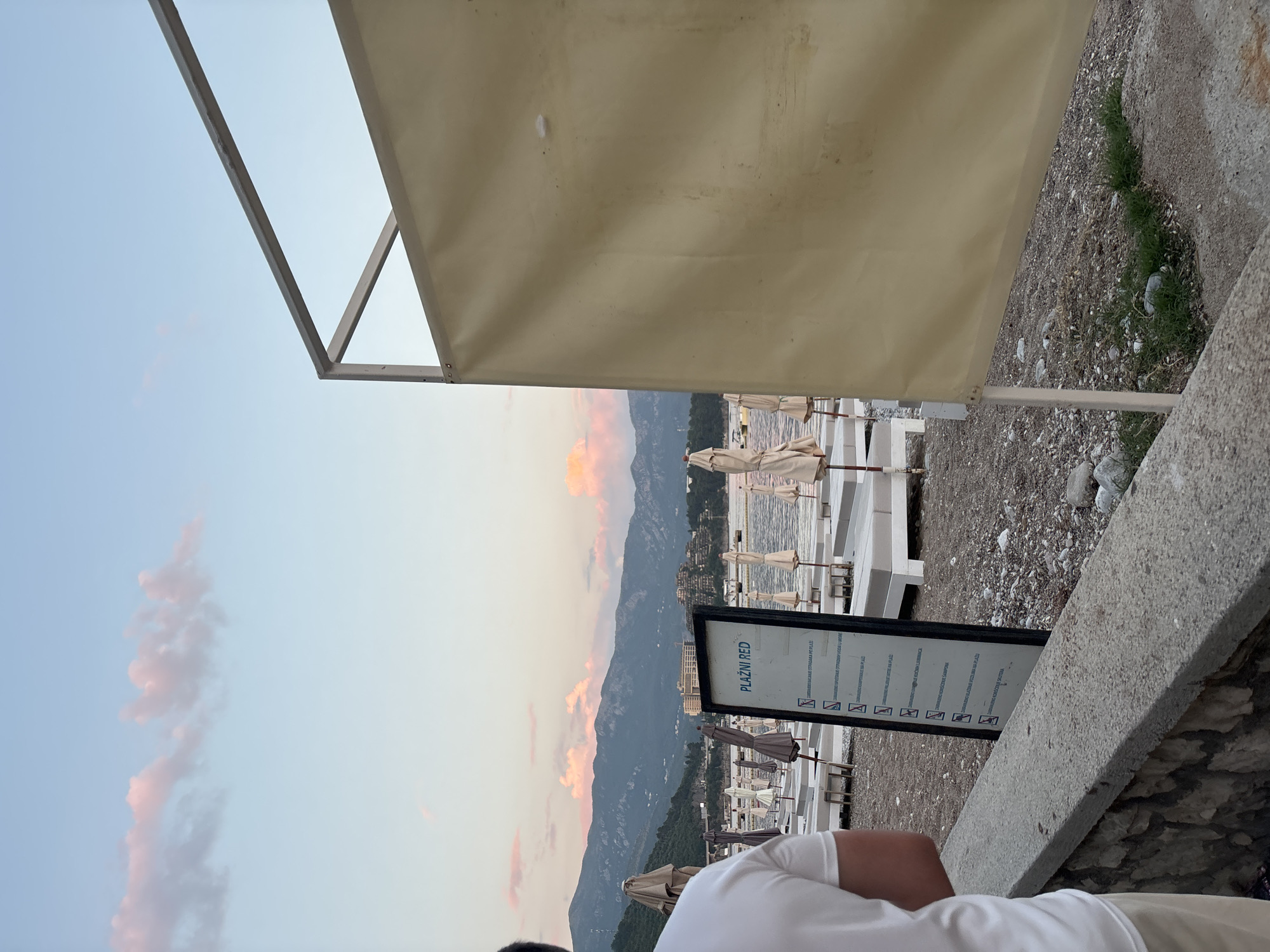 The image shows a beach in Budva, Montenegro, at what appears to be dusk or dawn. The sky has streaks of pink clouds. The scene is taken from a slightly elevated position, possibly from a patio or balcony. There are rows of beach umbrellas and lounge chairs on the pebble beach below. A large shade awning dominates the upper portion of the picture. A sign with the text "PLAZNI RED" is visible near the foreground. A person wearing a white shirt is partially visible in the lower part of the frame, likely holding the camera. The background features a mountain range.