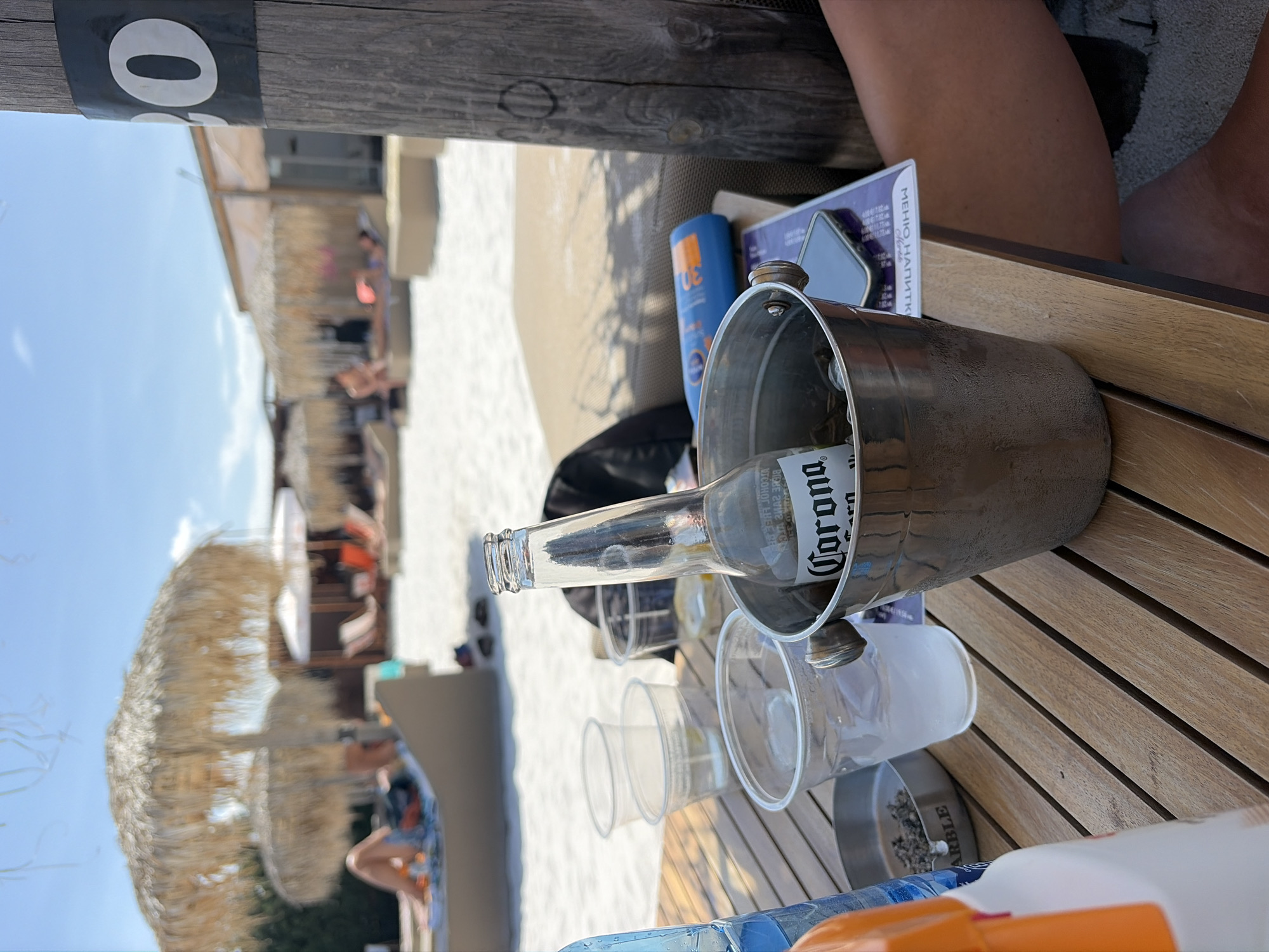 The image depicts a close-up view of a table at a beachside establishment, likely in Kazichene, Bulgaria, given the context of the prompt. The table, made of wooden slats, holds several items. Prominently featured is a silver ice bucket containing a partially full bottle of Corona beer and ice cubes. Next to it, there are three clear plastic cups, one containing a white cloudy liquid and ice, another empty, and a third with a small amount of clear liquid and ice. To the left, an ashtray filled with cigarette butts sits on the table. A blue bottle of what appears to be sunscreen is placed in the background, near a mobile phone and a menu. In the blurred background, beach umbrellas with thatched roofs and lounge chairs can be seen, with people relaxing on the sand under the bright daylight. A wooden post with the number "30" in a white circle on a black band is visible on the right side of the frame, suggesting a numbered seating area or section. A person's legs are partially visible in the lower right corner of the image. The overall scene suggests a casual, outdoor dining or lounging experience at a beach resort.
