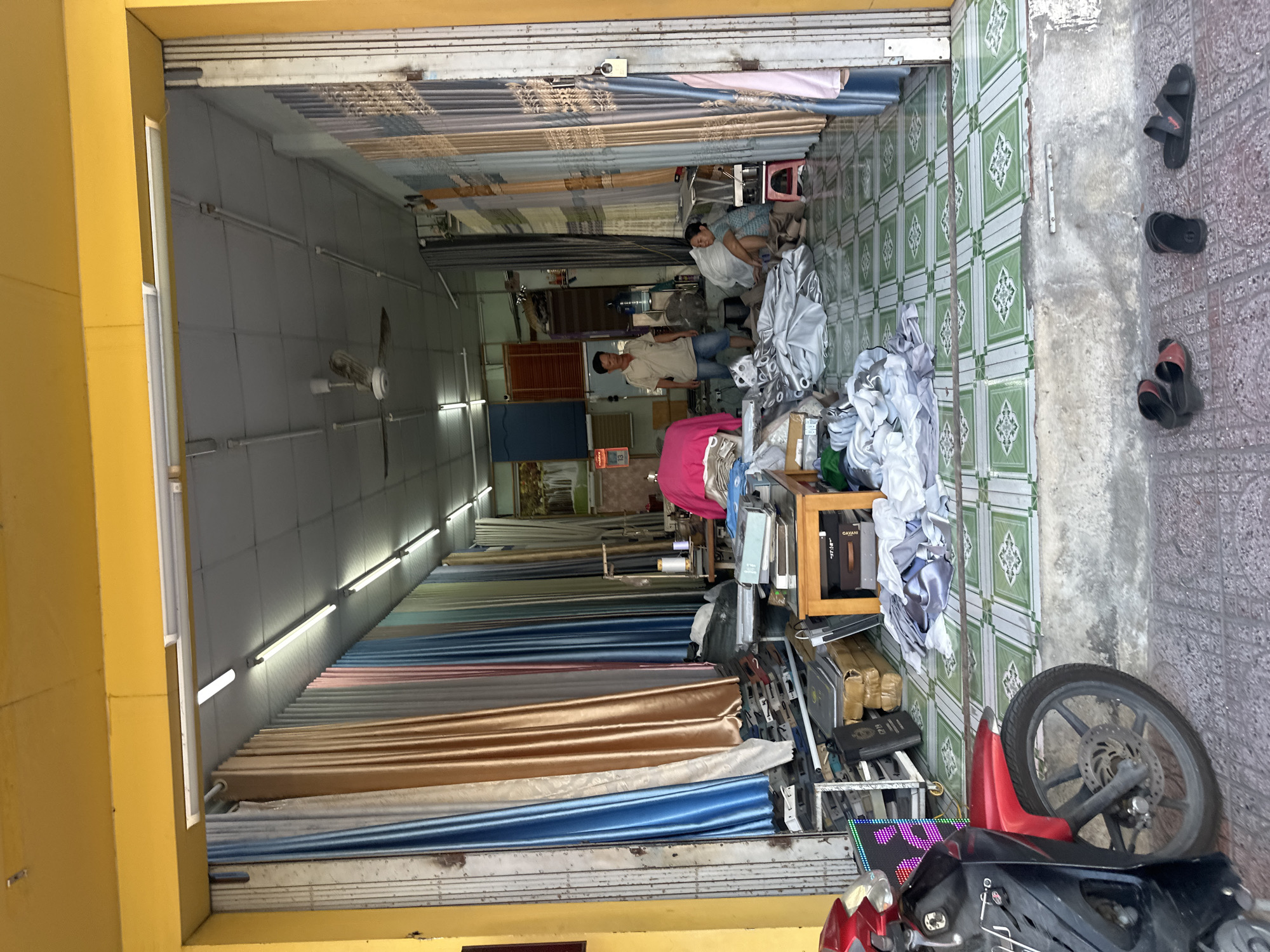 The image depicts the exterior and interior of a shop located in Bac Nha Trang Ward, Vietnam, specializing in textiles, likely curtains. The shop front is open, revealing a long, narrow space filled with rolled and hanging fabrics in various colors, including blues, browns, grays, and pastels. The floor is tiled with a green and white geometric pattern.
Inside, two individuals are visible. A man, wearing a light-colored polo shirt and blue shorts, stands near the center of the shop, looking towards the front. A woman, wearing a blue and white striped shirt and dark shorts, is seated on the floor further inside, appearing to be engaged with some fabric. Various items are scattered around the shop, including rolls of fabric, stacks of what appear to be books or fabric samples, a sewing machine, and a fan. A motorcycle is parked to the left of the entrance, its front wheel and handlebars visible. Several pairs of sandals are placed on the tiled ground outside the shop. The shop appears to be dimly lit, with fluorescent lights visible on the ceiling.