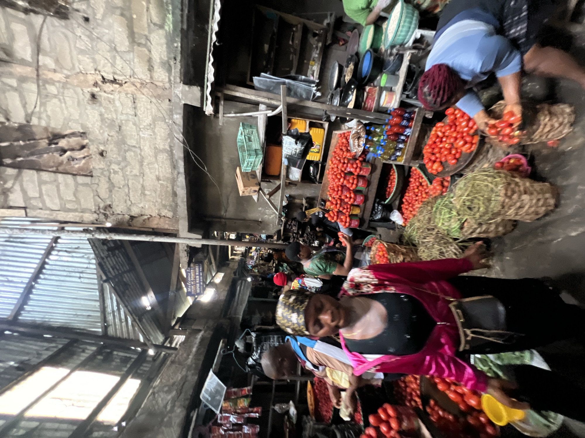 The image depicts a bustling outdoor market scene in Nkpor, Nigeria, likely a tomato market. A woman in the foreground, wearing a bright pink jacket over a black top and a patterned headscarf, is the central focus. She holds a yellow bucket in her left hand and her right hand is obscured. Behind her, a man wearing a brown and blue striped shirt is visible, looking to his right. To the right of the central woman, another woman in a blue shirt is actively sorting tomatoes from a woven basket into another. Numerous baskets filled with tomatoes are arranged on the ground and on stalls. The market stalls are constructed from rough, weathered materials, with corrugated metal roofing providing shelter. In the background, shelves are stocked with various goods, including bottles of what appear to be cooking oil and sauces, and crates containing produce. A sign in the upper background reads "Welcome to Jesus is Power Ltd. Nkpor Main Market." The overall environment is one of vibrant commercial activity, with people engaged in selling and buying produce under a makeshift, somewhat dilapidated structure.