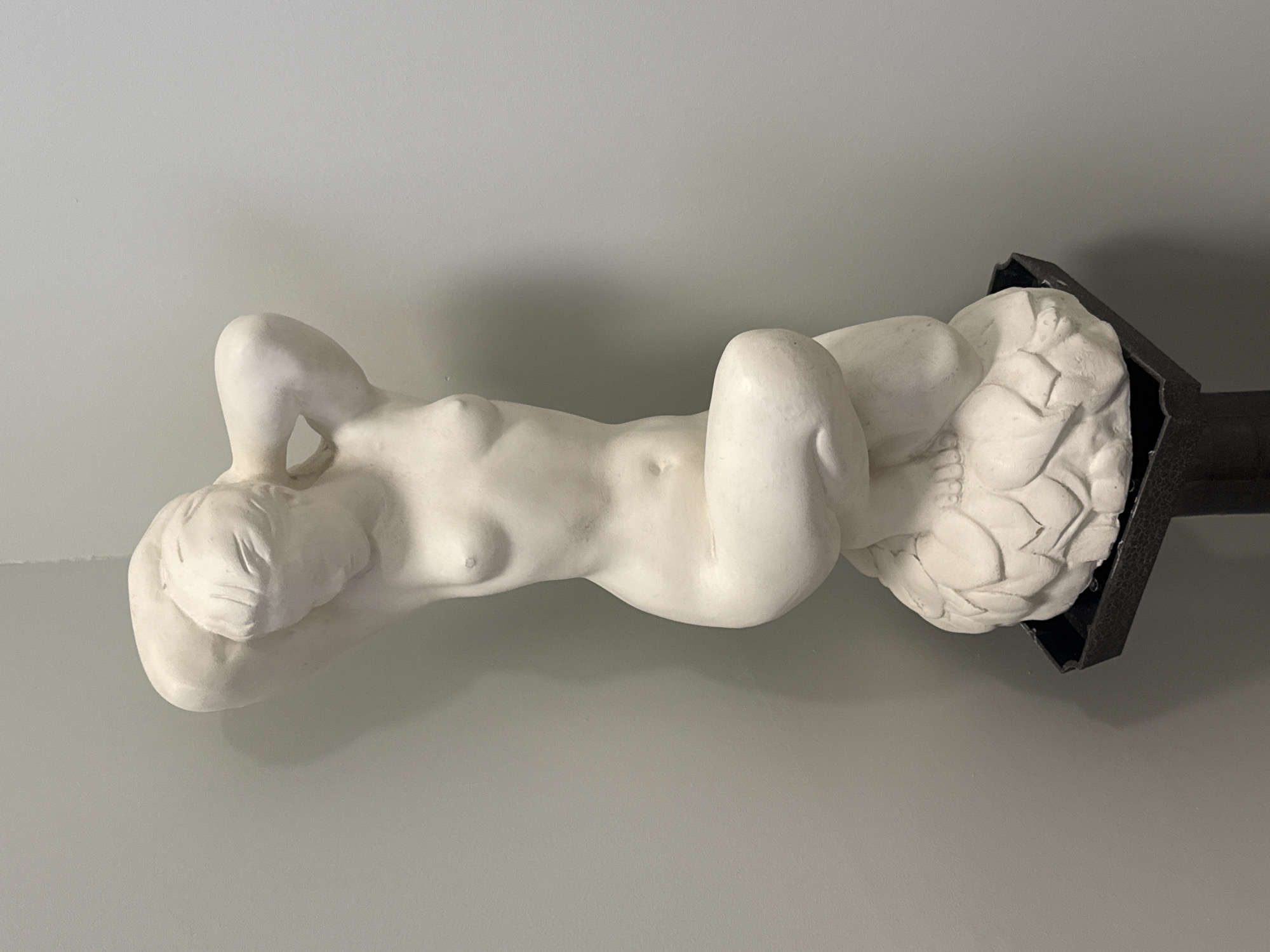 The image displays a white statue of a reclining nude female figure against a plain, light grey wall. The figure is made of a matte, light-colored material, likely plaster or ceramic, and exhibits a classical or neo-classical artistic style.
The statue depicts a woman lying on her back, with her head turned slightly to the left, resting on her bent left arm, which is positioned behind her head. Her right arm is bent, with the hand placed near her lower abdomen/thigh area. Her legs are drawn up, bent at the knees. The forms of the body are smooth and idealized, with subtle details like breasts and navel visible. The figure's lower body appears to rest on a dark, possibly black or very dark grey, structured base. This base has decorative, textured elements on its visible right side, resembling carved leaves or scales, specifically underneath where the statue's feet would be.
The setting appears to be indoors, indicated by the flat, uniform light grey wall serving as a background. Soft, diffused lighting illuminates the statue from an unseen source, casting gentle shadows on the wall behind and beneath the figure, adding depth to the scene. There is no discernible activity or event taking place, as the image captures a static sculpture. No visible text is present in the image. While the provided context suggests Saint Petersburg, Russia, there are no specific visual cues within the image to confirm this location.