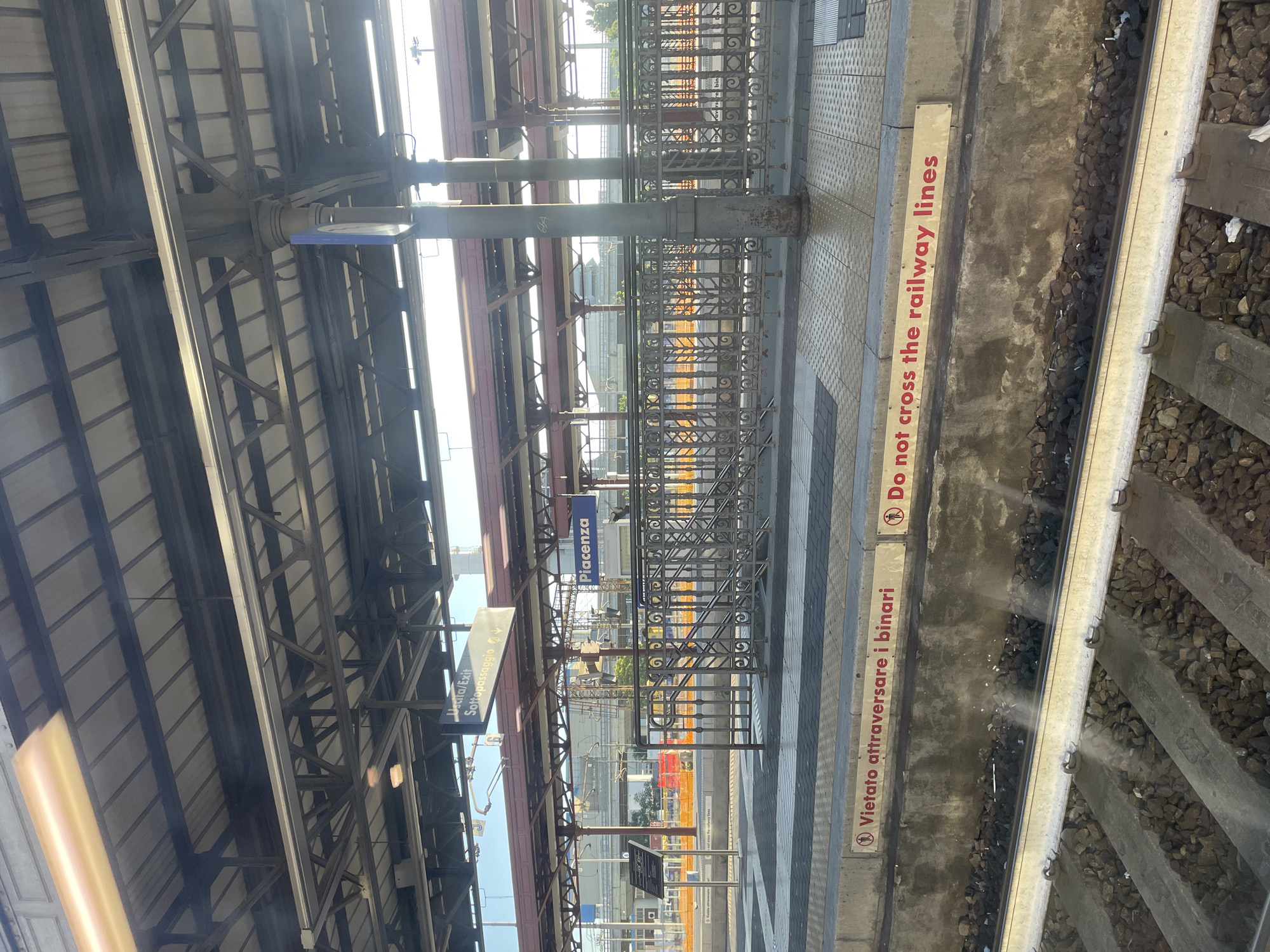 The image depicts the interior of a train station in Piacenza, Italy. Signage indicates "Uscira/Exit Sottopassaggio" and "Piacenza". A warning sign reads "Vietato attraversare i binari" and "Do not cross the railway lines". The image shows the train tracks and the edge of a platform with a metal fence. The station's roof structure is visible in the background.