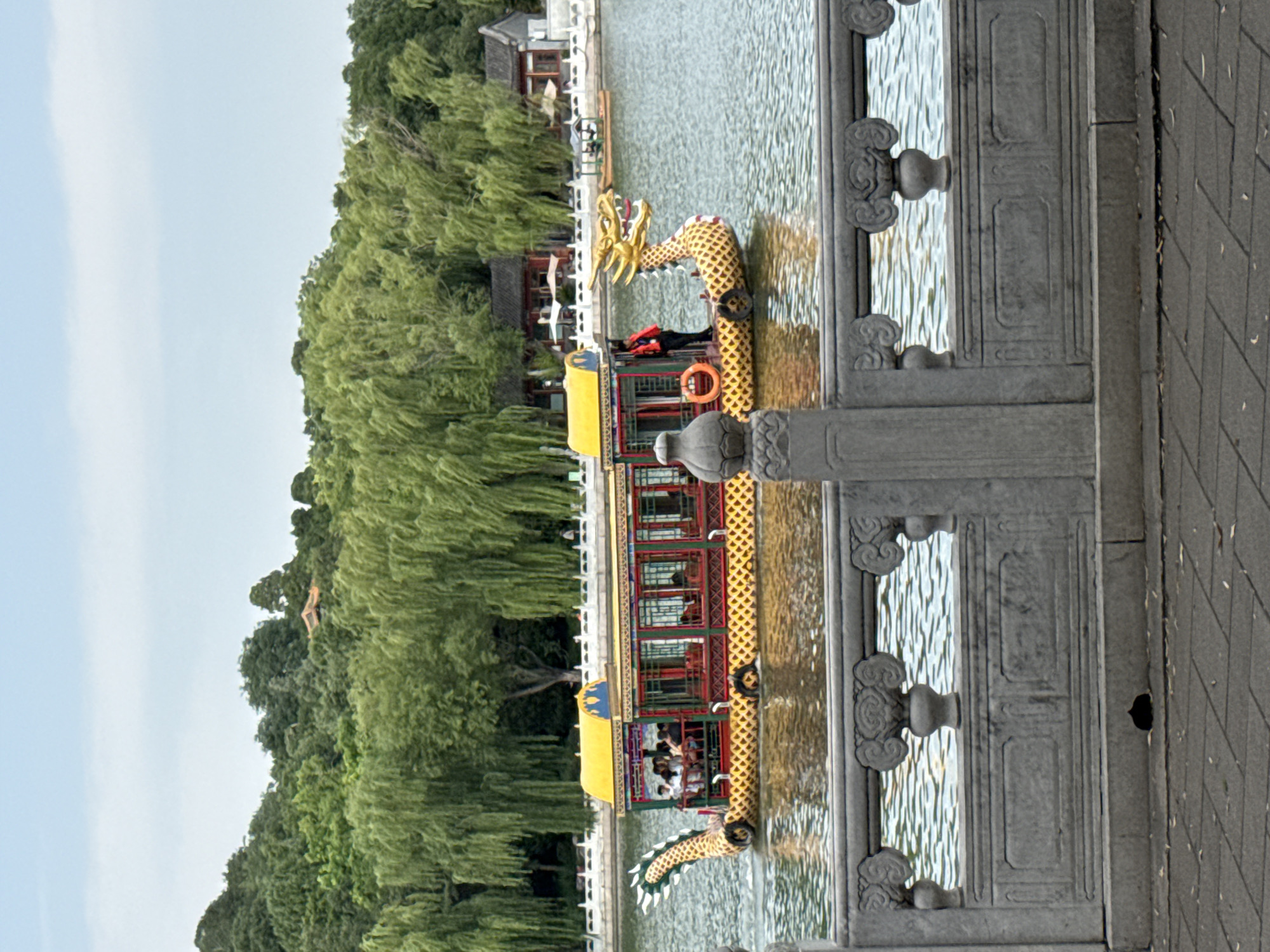 A traditional Chinese dragon boat carrying passengers glides across a calm body of water. The boat is ornately decorated with yellow, red, and green colors, and features a golden dragon head at its prow. Lush green trees and traditional Chinese architecture line the far bank of the water. In the foreground, a stone balustrade with decorative carvings separates the viewer from the water. The sky is a pale blue with some clouds. The scene appears to be set in a park or scenic area in Xicheng District, China, likely a place with historical significance and natural beauty.