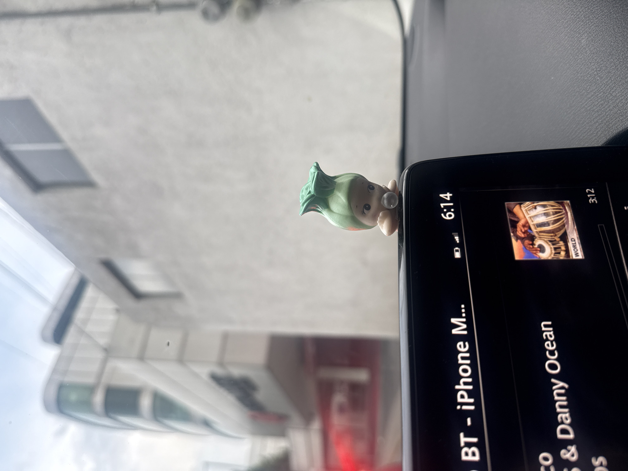 The image depicts the interior of a vehicle, likely a car, with a dashboard visible in the foreground. On the dashboard sits a black smartphone displaying a music interface, likely connected via Bluetooth, playing "Nico & Danny Ocean" under the "World" category with album art depicting a tabla. A small, whimsical toy figure with green hair and bubble gum is attached to the phone. Through the windshield, an out-of-focus view of a building exterior is seen, seemingly in Puebla, Mexico, based on architectural styles and urban layout. The time displayed on the phone is 6:14.