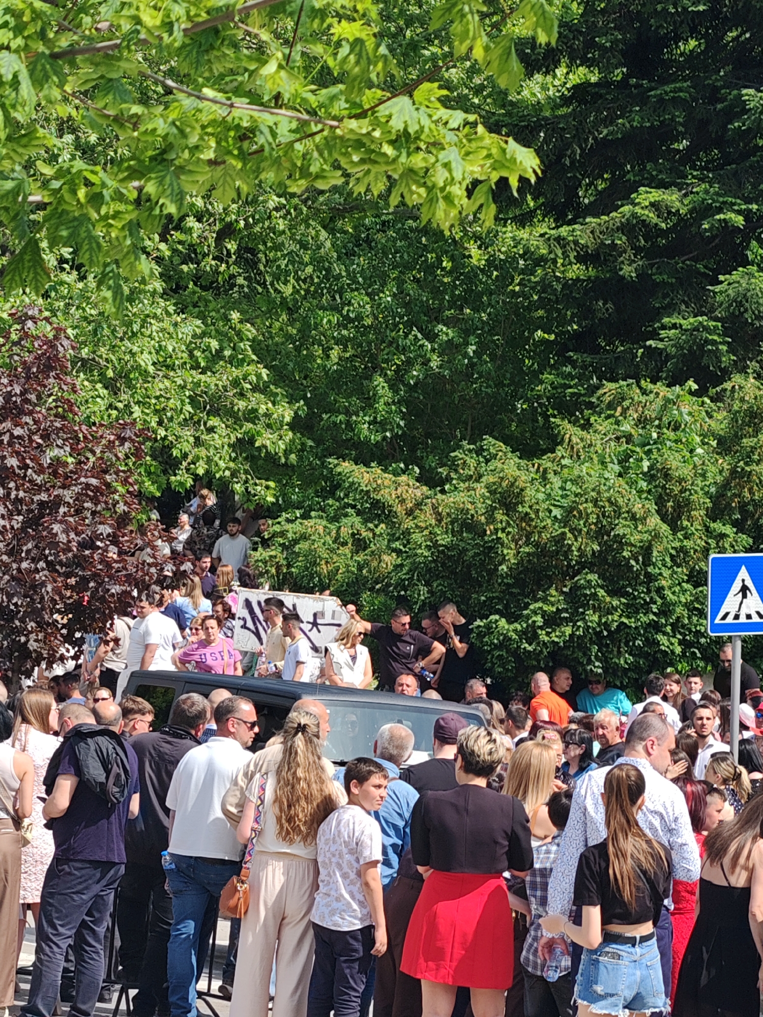 A large crowd of people gathers near dense green foliage in what appears to be an outdoor public space in Shumen, Bulgaria. The crowd surrounds a black vehicle, some individuals standing on top of it. A pedestrian crossing sign is visible on the right side of the frame. The gathering includes men, women, and children dressed in casual summer attire. The crowd's interaction or activity cannot be definitively determined, but it appears they are observing something or someone.