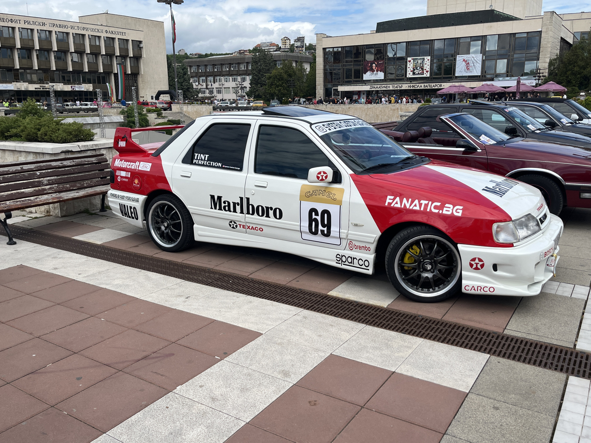 The image shows a white Ford Sierra modified for racing with red accents and sponsor logos like "Marlboro," "Texaco," and "Camel" with the number "69" displayed on the side. It features black wheels with yellow brake calipers. The car is parked on a patterned pavement near a metal grate and a wooden bench. In the background are several buildings, including one labeled "НЕОФИТ РИЛСКИ ПРАВНО-ИСТОРИЧЕСКИ ФАКУЛТЕТ" (Neofit Rilski Faculty of Law and History) and another building labeled "ДРАМАТИЧЕН ТЕАТЪР НИКОЛА ВАПЦАРОВ" (Drama Theater Nikola Vaptsarov) with people gathered near it. The overall setting suggests a public square or event location in the city of Unknown, Unknown. Several other cars are visible parked behind the racing car.