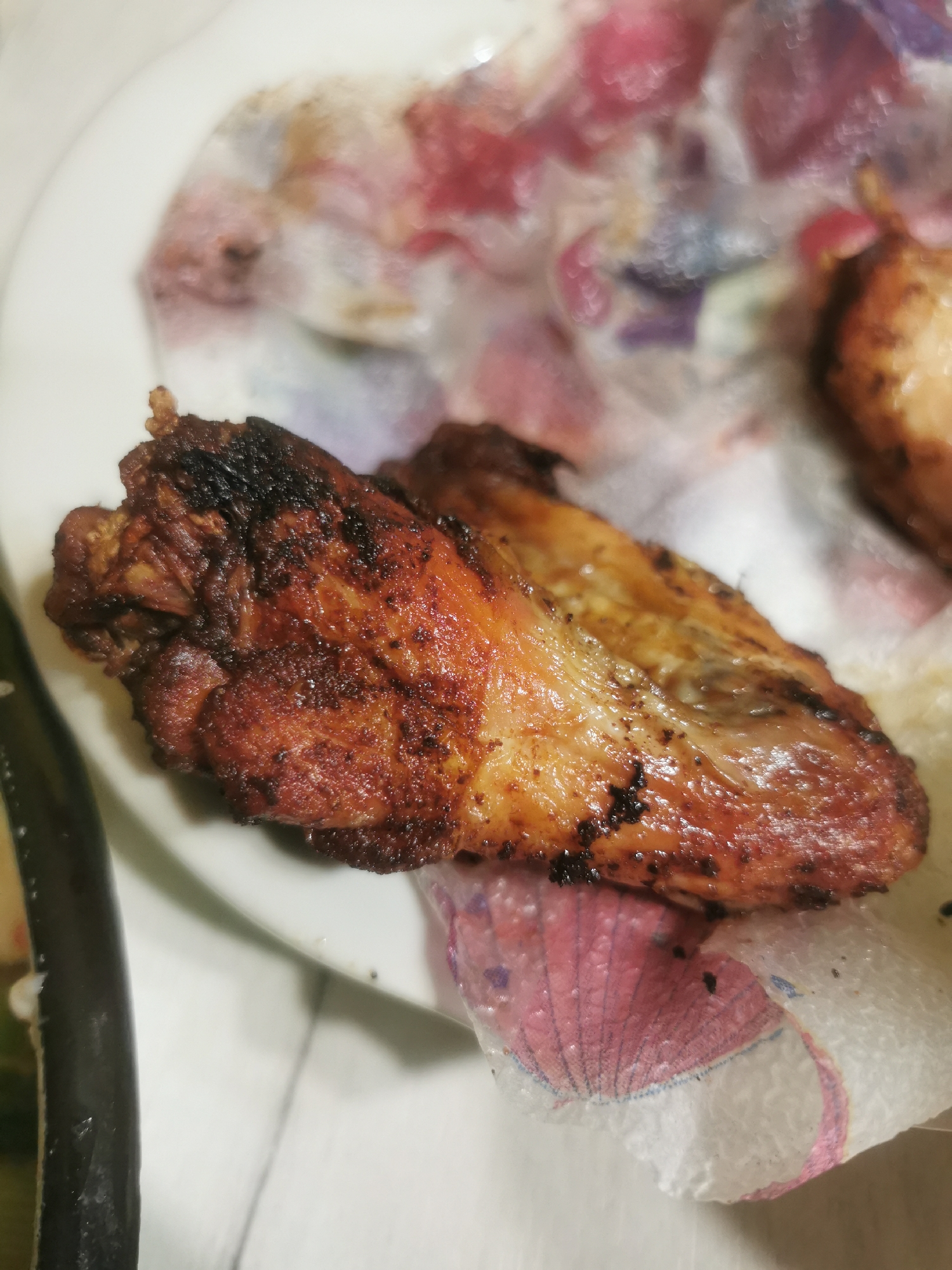 A close-up photograph shows a cooked chicken wing resting on a white plate with a decorative paper doily underneath it. Part of another piece of chicken is visible in the upper right corner. A dark, elongated object is partially visible in the lower left corner of the frame. The chicken appears well-done with visible dark spots, likely from grilling or frying. The doily features a pattern of pink and white. The overall scene appears to be indoors, likely in a domestic setting where food is being served. There is no indication of the location in Stara Zagora, Bulgaria.