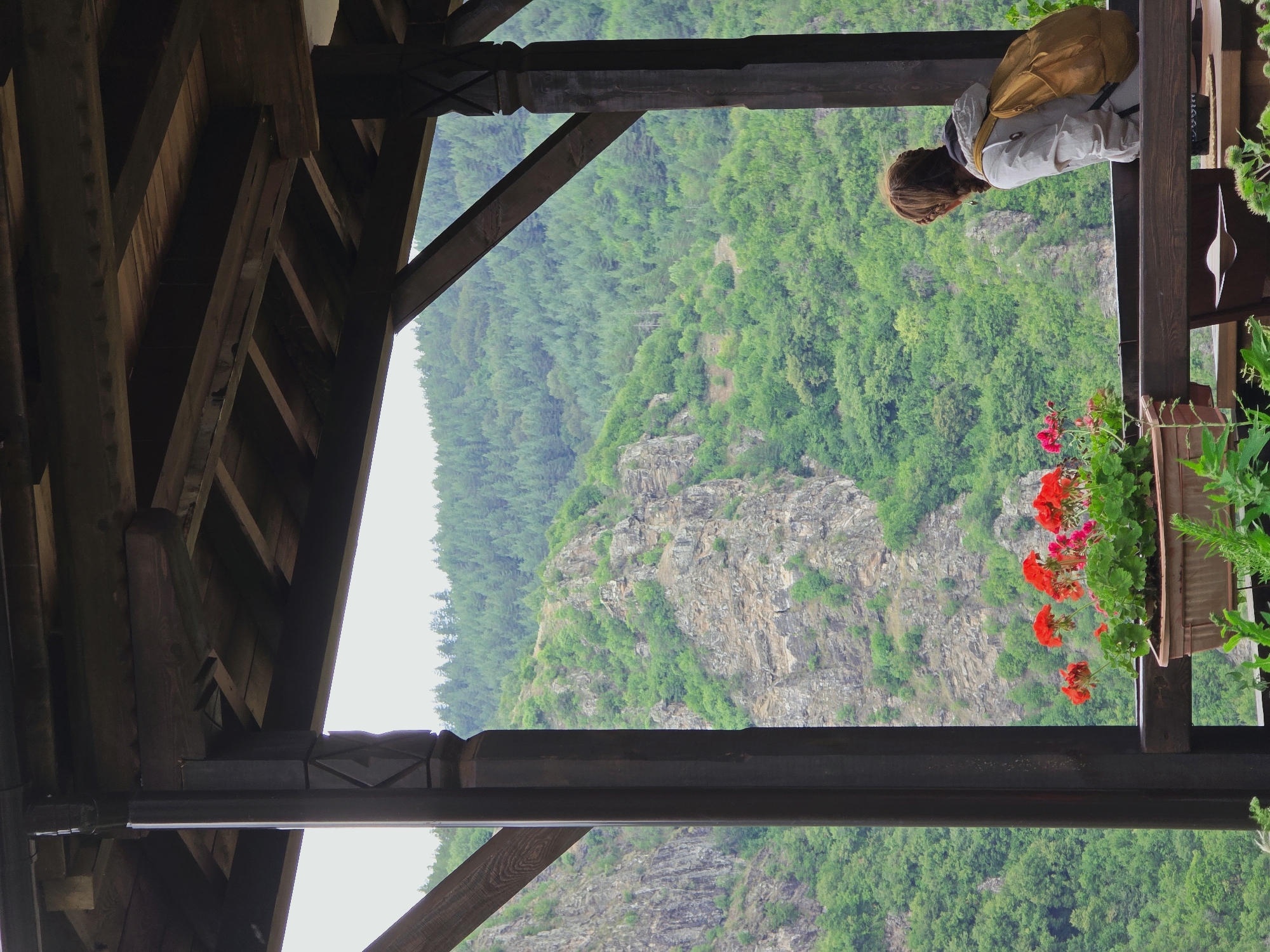 The image depicts a person with a backpack standing on a wooden balcony, overlooking a lush green mountainous landscape. The balcony has dark wooden pillars and a wooden ceiling. In the foreground, a planter box with red flowers is visible. The person, seen from behind, is wearing a light-colored jacket and is looking out at the scenery. The mountains in the background are covered with dense trees and rocky outcrops. The location is identified as Kovachevitsa, Bulgaria.