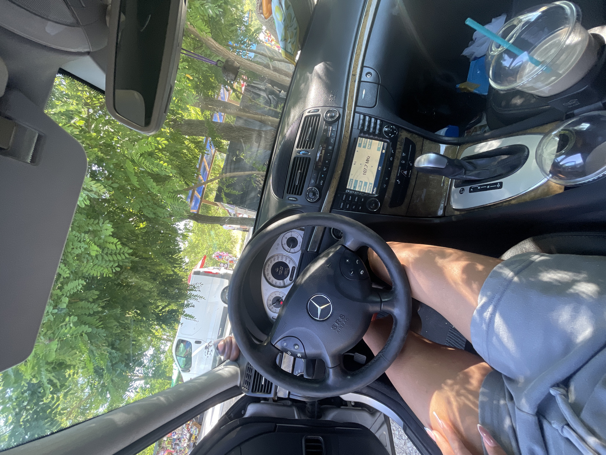The image presents a detailed view from inside a Mercedes-Benz vehicle, capturing both the interior cabin and the external environment on a sunny day in General Todorov, Bulgaria.
The lower half of a person, identified by tanned legs wearing light grey shorts, occupies what appears to be the driver's seat. One hand, featuring long, light-colored acrylic nails, rests on the left thigh.
The car's interior features a black Mercedes-Benz steering wheel with integrated controls and an "SRS AIRBAG" emblem. The instrument cluster displays a speedometer reaching 260 km/h, and a digital readout indicates an external temperature of 29.5°C. A small, brown, spiral-shaped novelty item is positioned on the dashboard. The center console includes an infotainment system showing "107.7 MHz" and "FM," along with climate control dials. An automatic gear shifter with a silver-trimmed base is prominently featured. Wood-grain accents are visible on the dashboard and console surfaces. Two cup holders are present in the center console, one containing a clear plastic cup with a light brown beverage and a teal straw. The driver's side door is ajar.
Outside the vehicle, lush green trees with abundant foliage create a dense canopy. Through the trees and the open door, a white panel van is partially visible. Further in the background, informal market stalls or roadside shops, characterized by blue tarpaulin roofs and various displayed items, suggest a commercial or gathering area. Strong, direct sunlight illuminates the scene, creating distinct shadows and bright highlights.