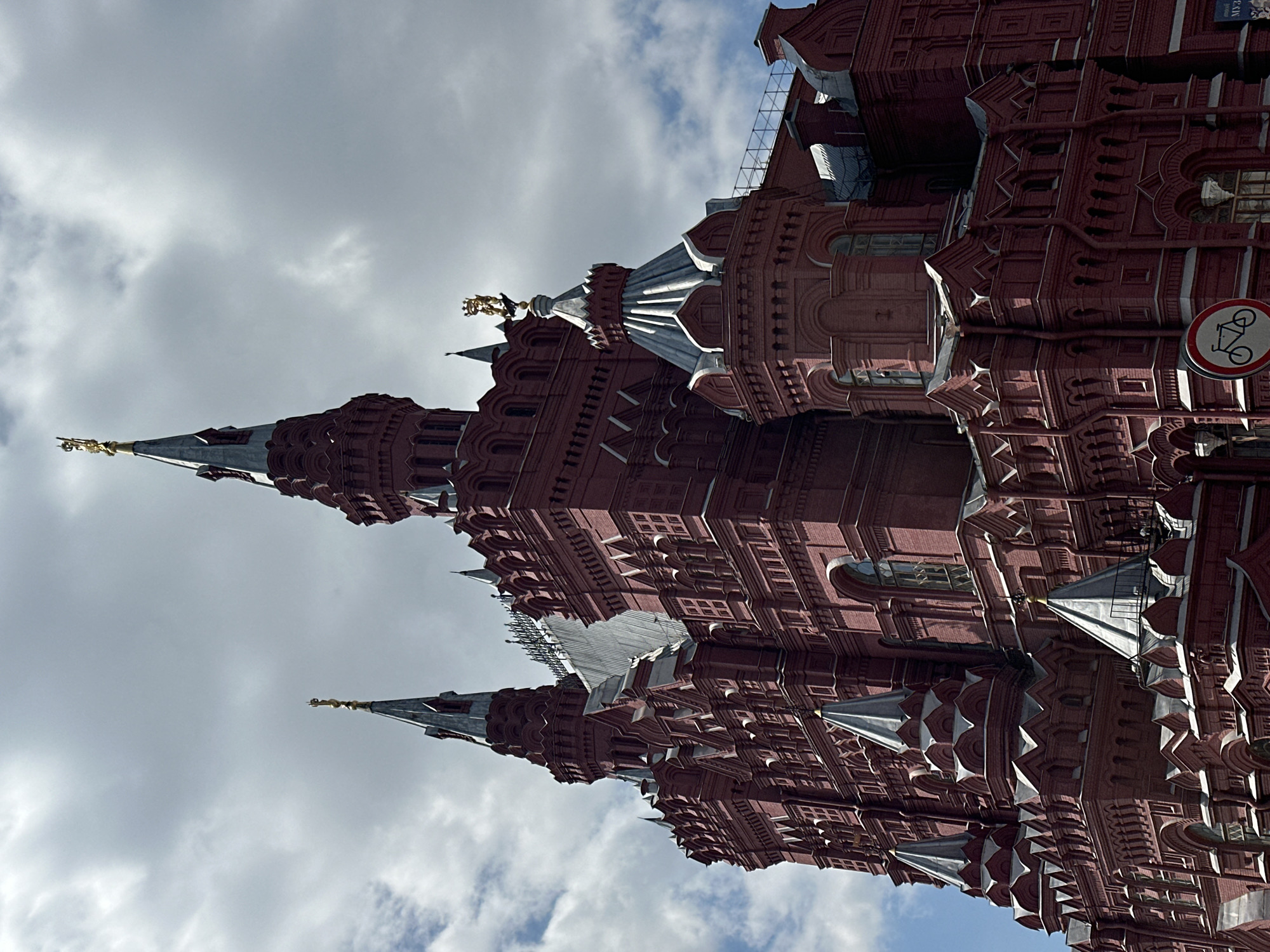 This is a medium-shot, exterior, eye-level, vertical photograph of the State Historical Museum in Moscow, Russia, viewed from a low angle looking up towards the sky. The building is constructed of red brick and features intricate architectural details, including numerous towers, spires, and decorative elements. The sky is cloudy with patches of blue visible. In the lower right corner of the image, a circular traffic sign with a bicycle symbol is visible, along with a partial view of the museum's name in Cyrillic. The lighting suggests it is daytime, with shadows indicating a partly cloudy sky. No people are present in the frame.