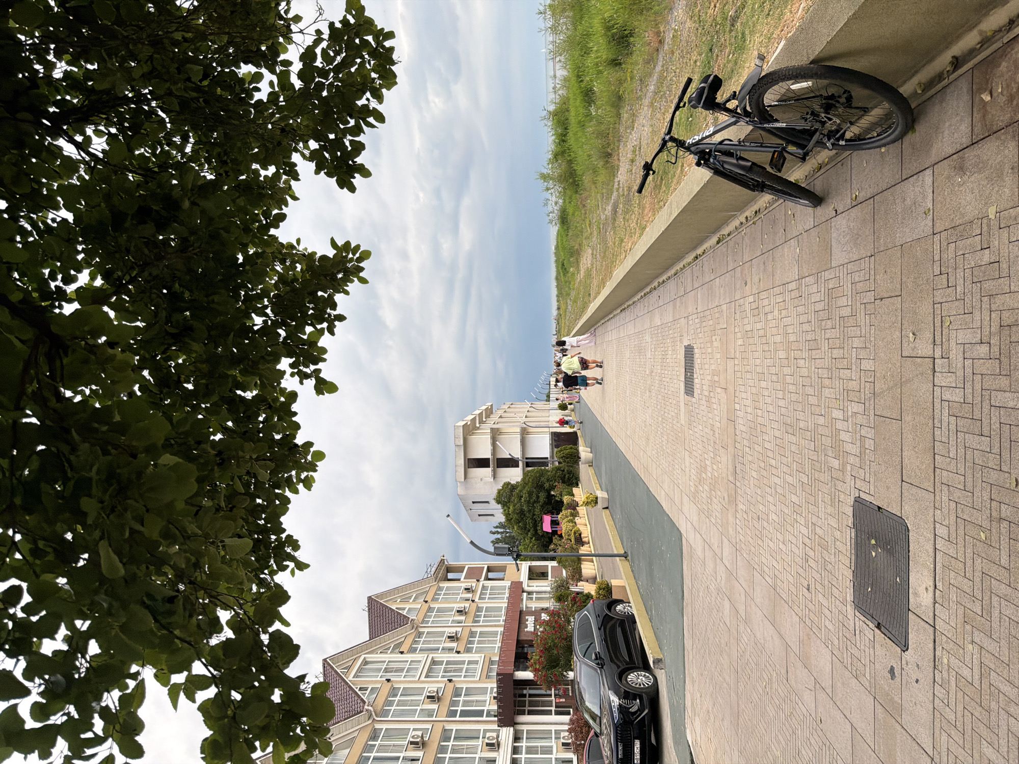 The image captures a scenic view of a pedestrian walkway, likely in Sochi, Russia, adjacent to a grassy embankment and the sea in the distance. The walkway is paved with light-colored bricks laid in a herringbone pattern. A black bicycle is parked along the concrete barrier separating the walkway from the embankment.
Several people are strolling along the walkway, dressed in casual summer attire, suggesting a pleasant day. In the background, modern buildings with multiple windows and balconies line the street, with one building displaying a sign that appears to read "Hotel". A black car is parked on the side of the road next to the buildings. Lush green trees, with dense foliage, frame the left side of the image, contributing to the natural beauty of the scene.
The sky is partly cloudy, indicating a mix of sun and clouds, typical of a daytime setting. The overall atmosphere appears relaxed and leisurely, suggesting a typical day in a coastal resort town. The presence of people walking, a parked bicycle, and buildings implies an urban or semi-urban coastal environment. No specific time of day is definitively indicated, but the lighting suggests it is daytime.