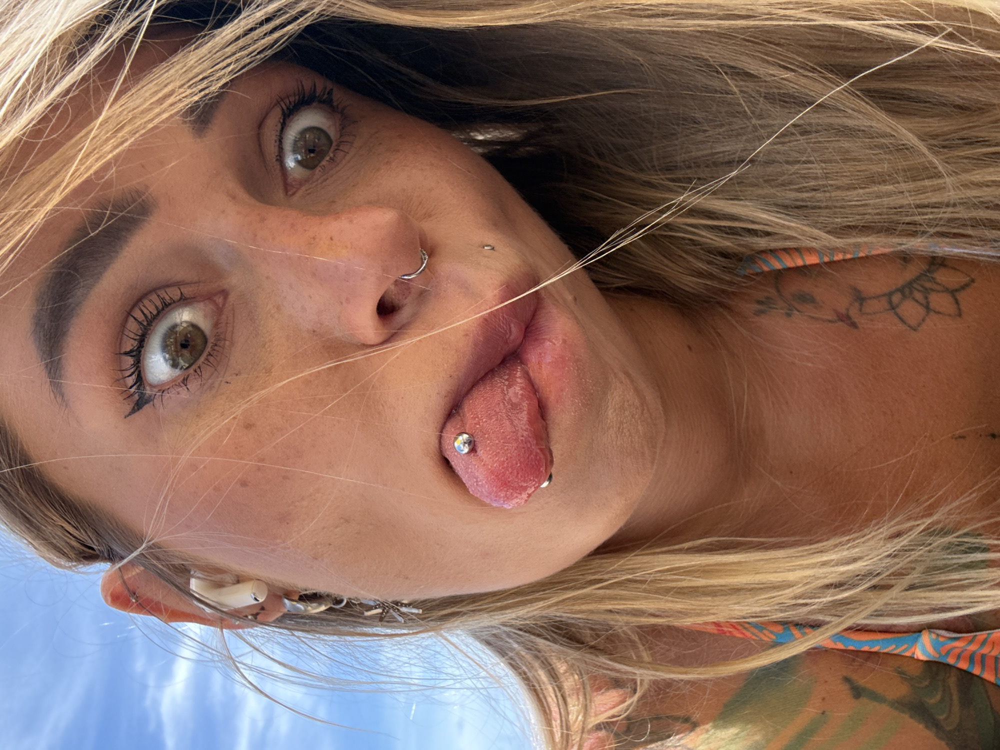 A close-up, low-angle shot captures a young Caucasian woman in Choeng Thale, Thailand, against a bright blue sky. She is looking directly at the camera with a playful expression, her eyes wide open, and her tongue sticking out, revealing a silver tongue piercing. Her mouth is slightly open, showing her teeth and the piercing. She has blonde hair, some of which falls across her face, and appears to be wearing a bikini top with an orange and blue pattern. She also has several ear piercings, one of which is a dangling earring. A discreet nose ring and a small piercing below her lower lip are also visible. The background is predominantly the sky, suggesting an outdoor setting.