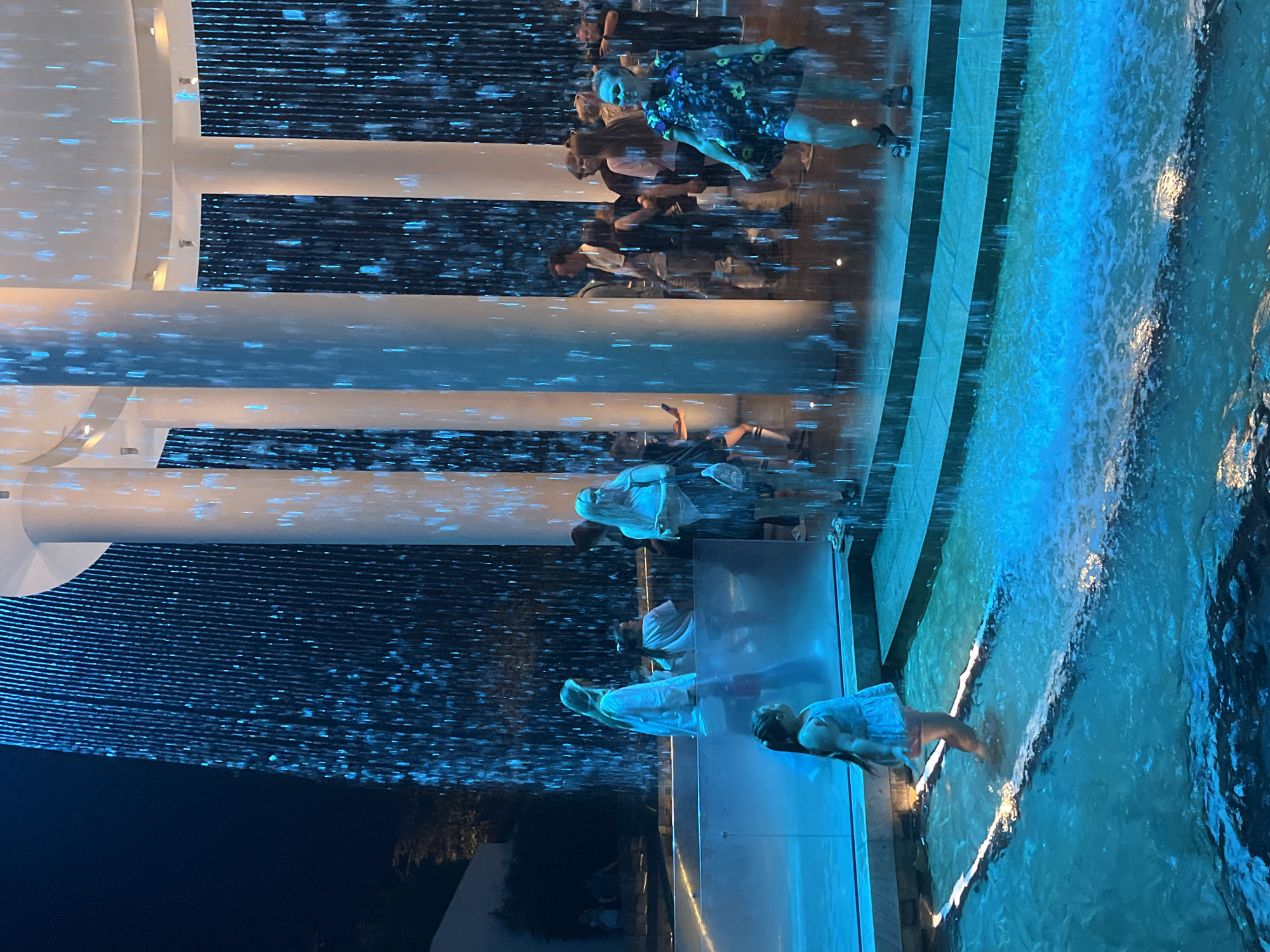 The image captures a modern architectural space with a dramatic waterfall feature. The waterfall, illuminated with blue light, cascades down behind several large, white columns. A group of people are visible, some standing near the waterfall and others in the water feature itself. In the foreground, a young girl in a light-colored dress is standing in the shallow water, her back to the camera, looking towards the waterfall. Other individuals, both adults and children, are scattered throughout the scene, interacting with the environment and each other. The overall impression is one of a public space designed for visual spectacle and visitor engagement. The question of this scene being located in Krasnodar, Russia, cannot be answered from the visual information provided in the image.