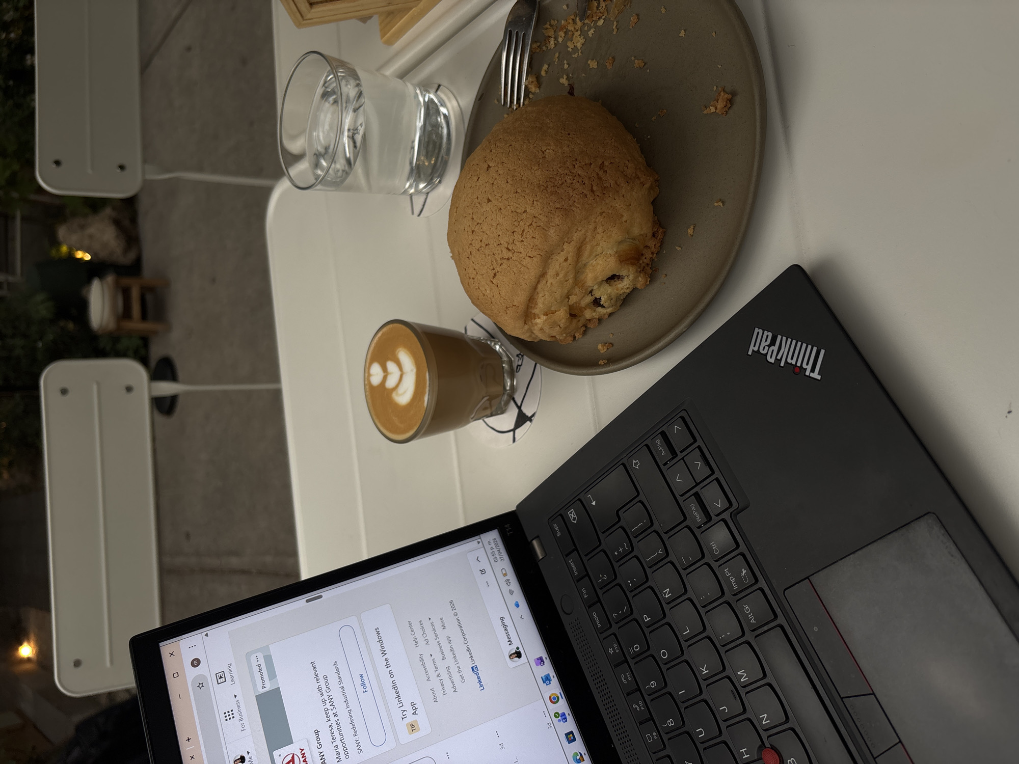 The image displays a serene outdoor cafe setting, likely in Mexico City as suggested by the context, during the late afternoon or early evening.
**Subjects & Objects:** The main subjects are a black ThinkPad laptop, a clear glass of water, and a glass of latte with a heart-shaped latte art. A large, round pastry, resembling a Mexican concha or pan dulce with a crispy topping, rests on a grey plate, accompanied by a fork. Crumbs on the plate indicate it has been partially consumed. These items are arranged on an off-white table. The background features other white outdoor furniture, such as chairs or tables, and some blurred greenery and warm ambient lights, hinting at an outdoor patio environment.
**Activity:** The scene captures a person engaged in work or online browsing on their laptop while enjoying a beverage and a sweet pastry. It suggests a casual work session or a relaxing break in an outdoor setting.
**Notable Details:** The time displayed on the laptop screen is "05:55 p.m." on "27/04/2026", which aligns with the soft, dimming light in the image and the presence of artificial background lights. The overall atmosphere appears calm and focused.
**Visible Text:** The laptop's lid clearly shows the "ThinkPad" logo. The screen displays a LinkedIn page with text elements such as "Promoted...", "ANY Group", "María Teresa, keep up with relevant opportunities at SANY Group", "SANY: Redefining Industrial Standards", "Follow", "TIP Try Linkedin on the Windows App", "About Accessibility Help Center Privacy & Terms Ad Choices Advertising Business Services Get the Linkedin app More Linkedin Linkedin Corporation © 2026", and "Messaging". Various keyboard keys are also visible, including "Ctrl", "Alt Gr", "Imp Pt", "Supr", "Insert", "Fin", "Inicio", "AvPág", and "RePag", alongside letter and number keys.