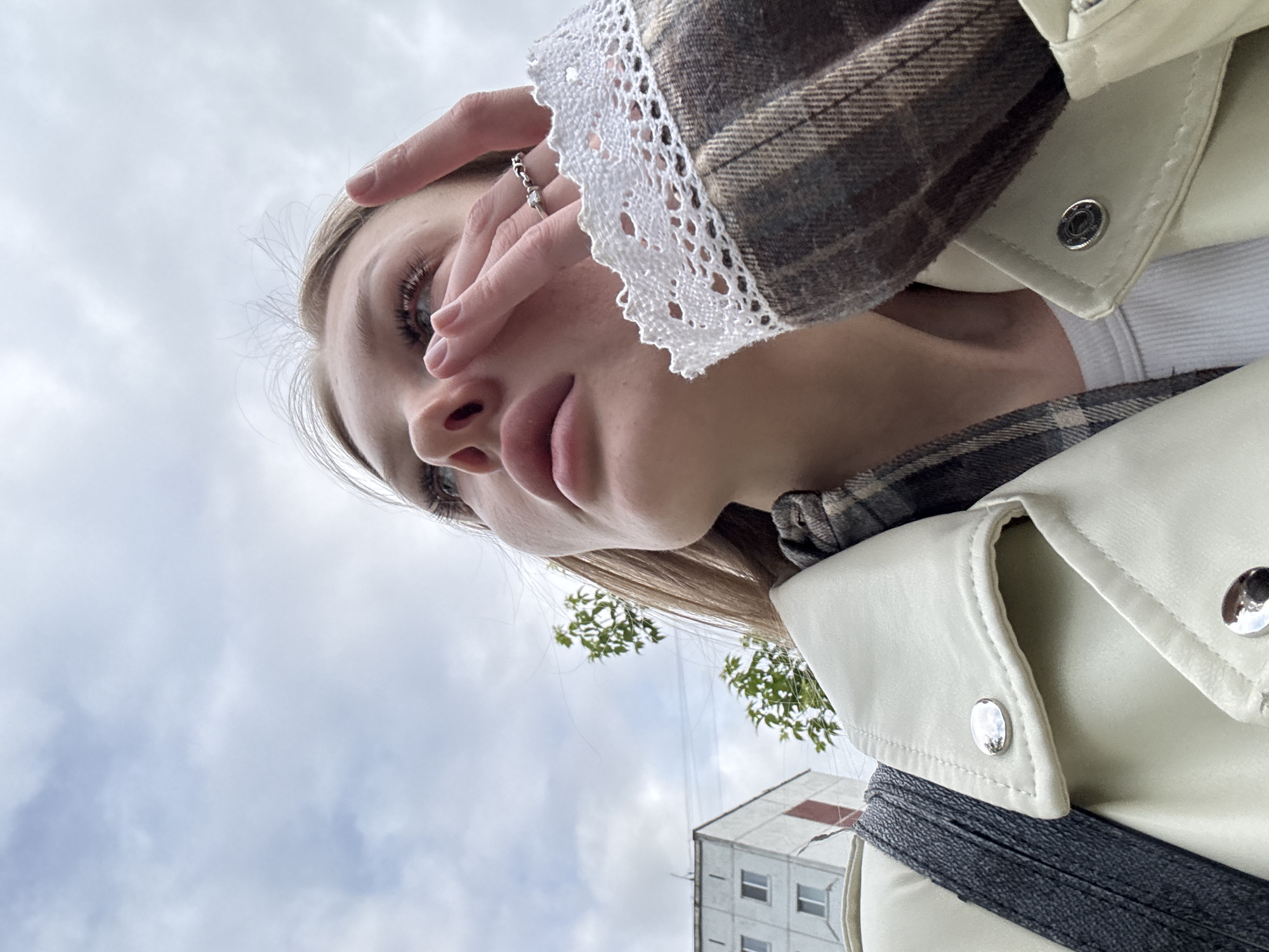 A low-angle portrait depicts a young woman. The woman has fair skin, blonde hair, and full lips. She is wearing a light-colored jacket with large metal buttons and a checkered shirt or jacket with a lace trim on the sleeve. Her hand, adorned with a ring, is partially covering her face, possibly shielding her eyes from the sun.
The background reveals a cloudy sky and a glimpse of an urban environment. The corner of a multi-story building with small windows is visible. Based on this information, it's impossible to confirm the exact location within Perm, Russia. More context would be needed to specify the area.