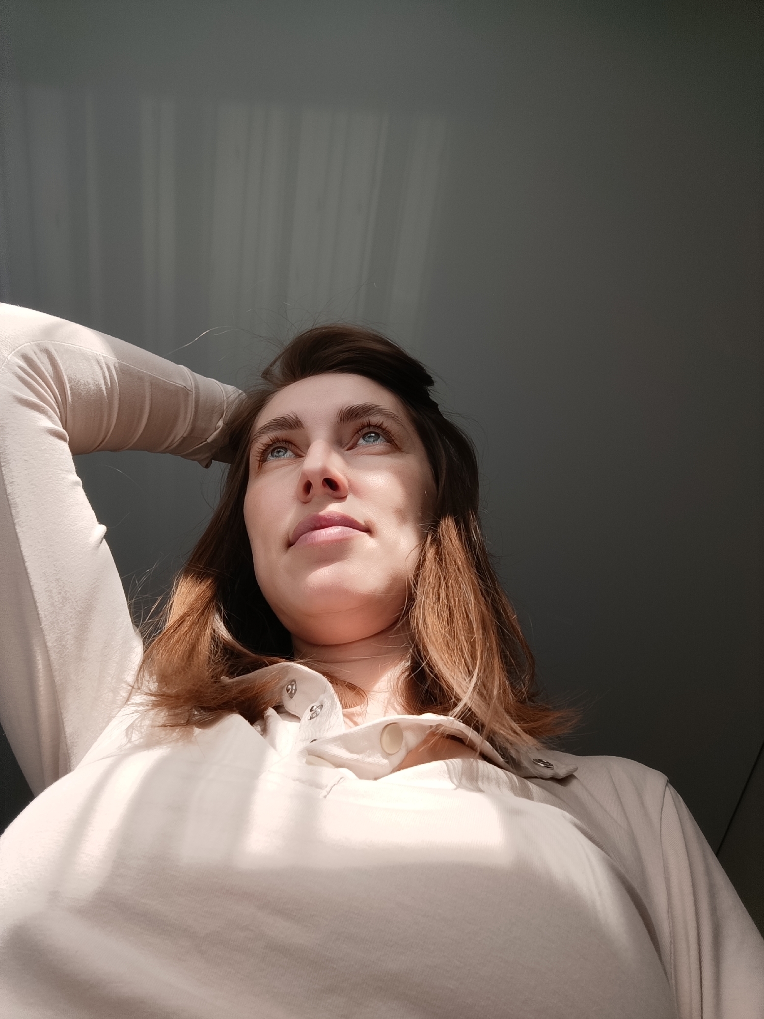 A woman with light skin, brown hair, and blue eyes is gazing upward. She is wearing a light-colored top with buttons at the collar. Her left arm is raised and her hand rests on her head. The background is a light gray wall with light filtering across it. The location cannot be determined from the image.