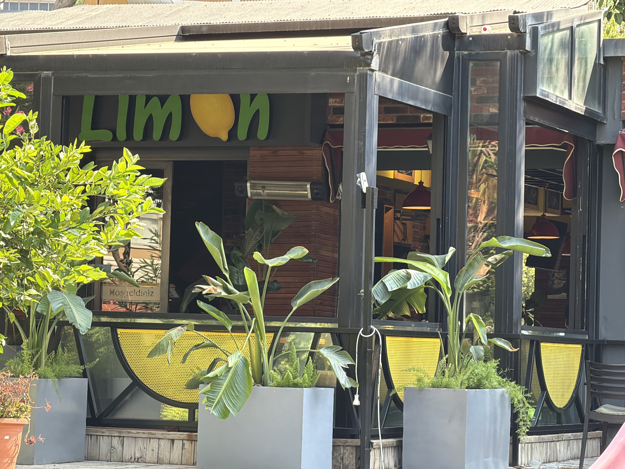 The image shows the exterior of a cafe named "Limon" in Alanya, Turkey. The cafe has a dark-colored facade with large windows and an outdoor seating area. A bright green "Limon" sign with a yellow lemon illustration is prominently displayed above the entrance. The cafe is decorated with several potted plants, including large banana-leaf plants and smaller green bushes. The outdoor seating area features wooden decking and a black metal chair. A sign that says "Hoşgeldiniz" (Welcome in Turkish) is visible through the window. The overall impression is a welcoming and lively atmosphere.