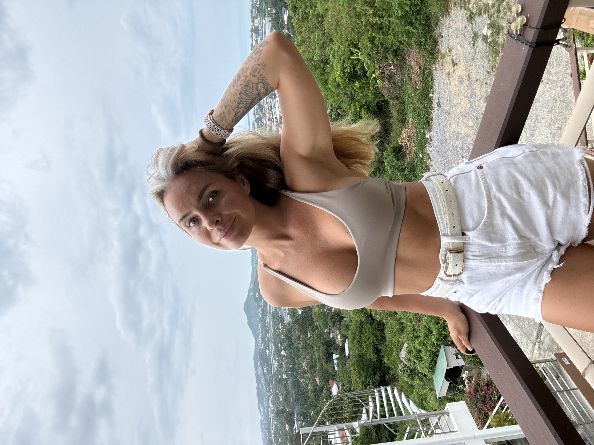 A blonde woman with tattoos on her arm is standing on a balcony overlooking a lush green landscape with a city and mountains in the distance. She is wearing a beige sports bra and white denim shorts with a white belt. The woman is looking at the camera with her hand on her head. The sky is overcast. The location is likely in the Ko Samui District of Thailand, given the tropical vegetation and architecture.