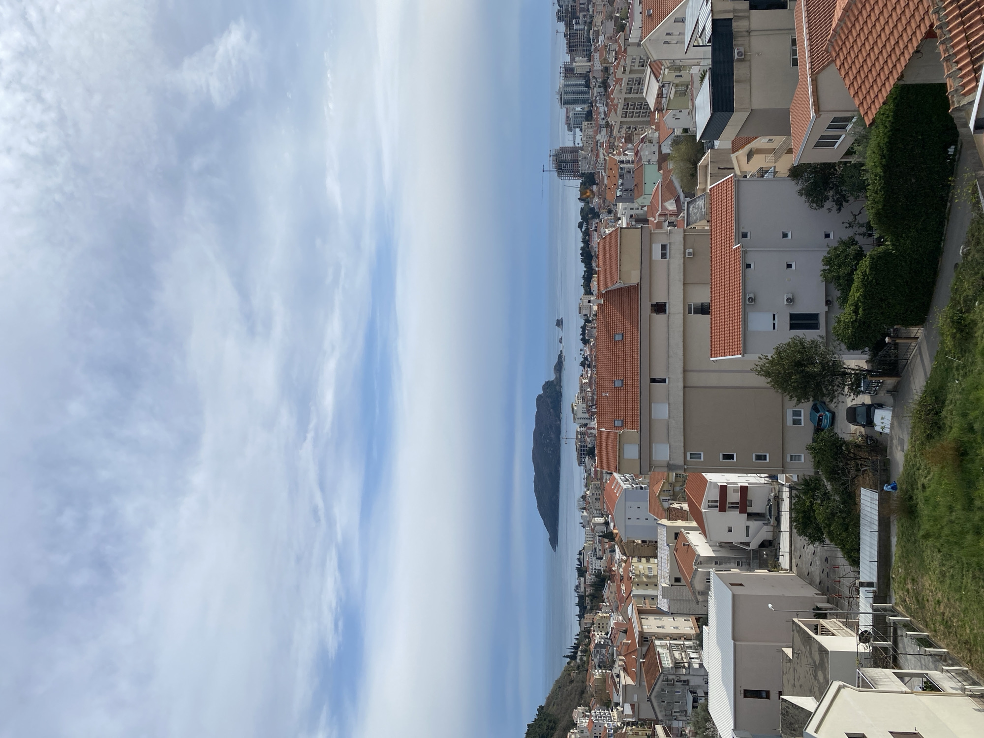 The image depicts a cityscape of Budva, Montenegro, from an elevated vantage point, likely a high-rise building or hillside. The scene is bisected by a view of the Adriatic Sea, with Sveti Nikola Island visible offshore. The sky is bright with patchy clouds. The urban landscape consists of densely packed buildings, primarily residential, with red-tiled roofs. Streets are visible but mostly obscured by the structures. A few parked cars are visible. Vegetation including trees and grass are also visible among the buildings. There are no people visible in the shot.