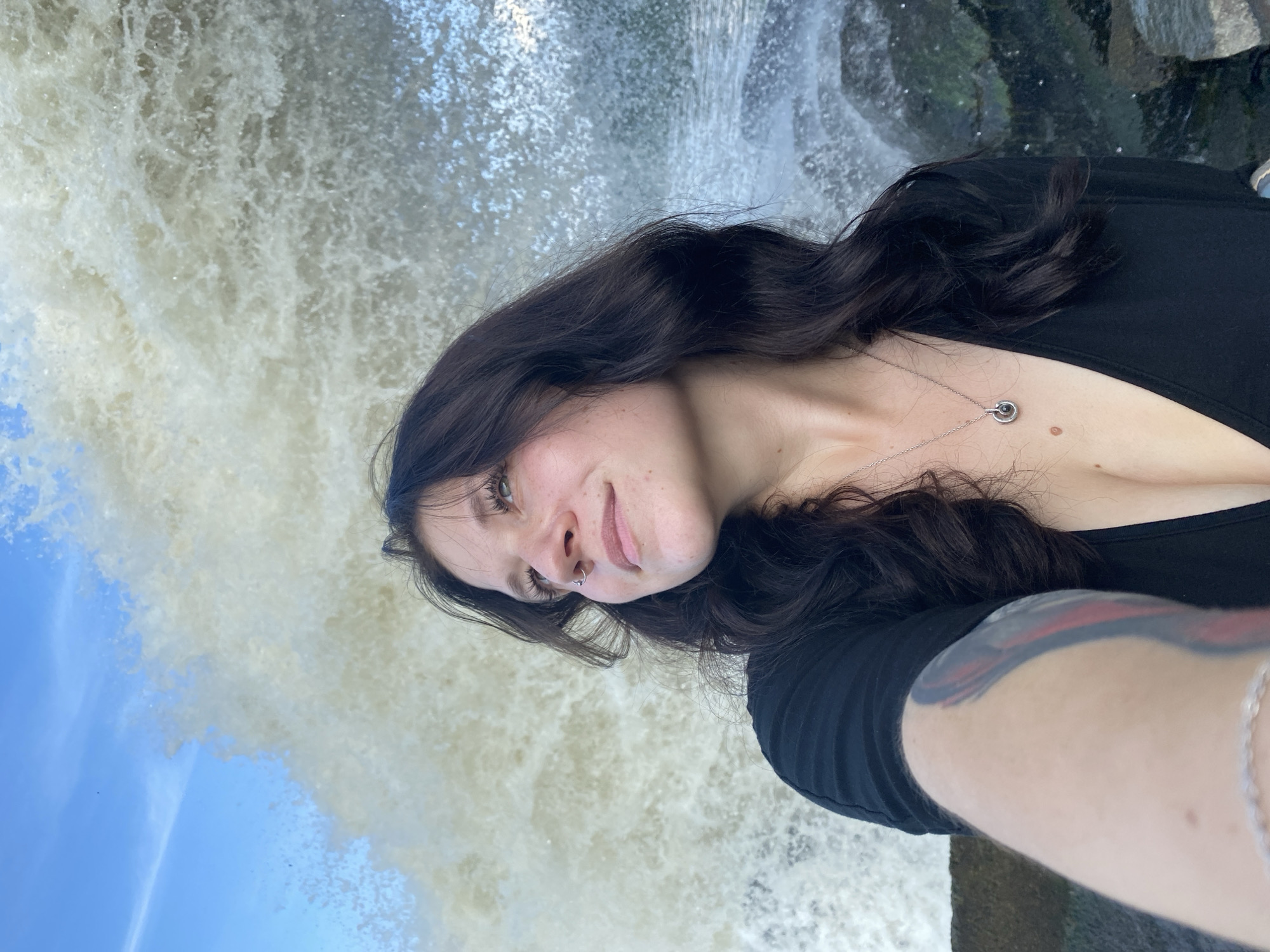 A young woman with dark hair and a nose ring poses for a selfie in front of a crashing wave. She is wearing a low-cut black top and a necklace. Her arm, which is visible in the foreground, has a tattoo. The background features a large wave breaking with white spray, and a blue sky with some clouds. The presence of a crashing wave suggests a coastal location, possibly on the shore of the Ural River, which flows through Chernushka, Russia.