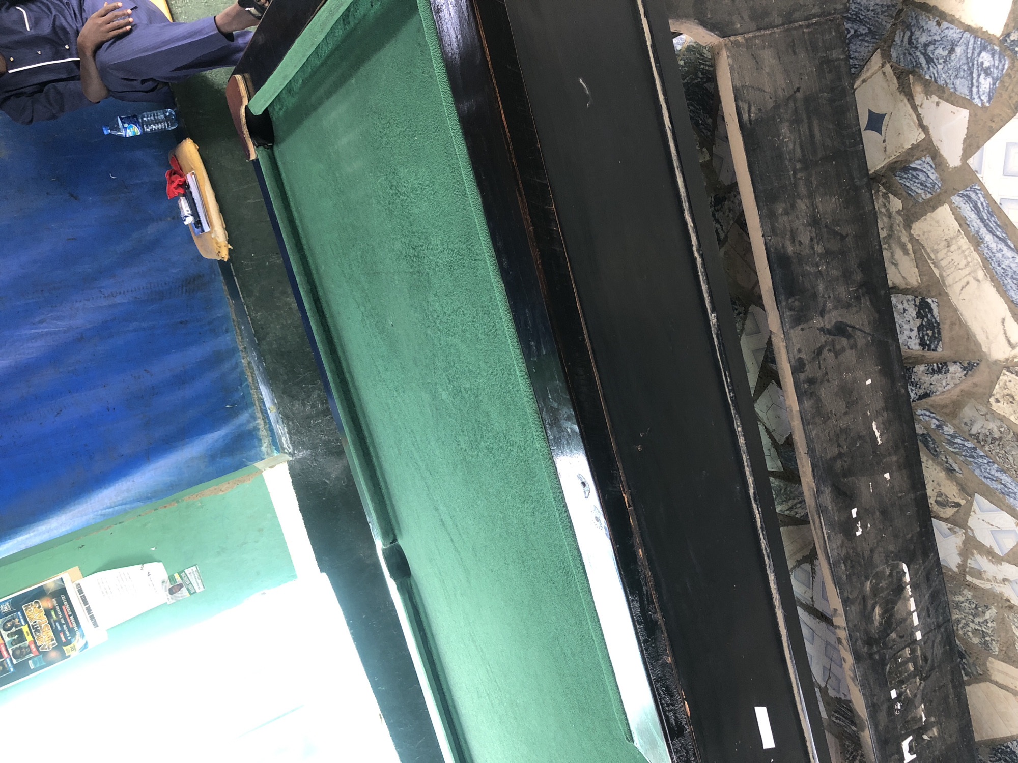 The image depicts an indoor scene within an Unknown City, Unknown Region, likely a pool hall or recreational area. The primary subject is a billiard table with a worn green felt surface and dark wooden rails. A white billiard cue is laid horizontally on the left rail, and a wooden triangle rack rests on the table's far right end. No billiard balls are visible on the table. The table's black wooden frame shows considerable wear, including scratches and chipped paint.
In the upper right quadrant, a single individual is partially visible, seated on a yellow cushion placed on a low ledge against a textured, dark blue wall. The person is dressed in a dark blue tunic with white vertical trim on the placket and matching dark trousers, wearing dark sandals. The individual appears to be resting or observing. Adjacent to the person on the ledge are a clear plastic water bottle with a blue label, a small red cloth, a metallic object, and a book or stack of papers.
The background to the left features a light green wall adorned with several paper items. A larger poster, partially obscured by overexposure, displays images of people and includes text fragments such as "NIGHT OUT" and "AMAZON POOL HOUSE." Below this poster are smaller white slips of paper and a small portrait of a man, with the name "MANN MANCHA" partially legible.
The floor exhibits an irregular mosaic pattern constructed from varying sizes and shapes of tiles in white, light blue, dark blue, beige, and grey, set with a light brownish-grey grout. The area beneath the billiard table's underframe appears dark and accumulated with dust. The overall environment suggests a well-used, somewhat aged public or semi-public recreational facility, characterized by utilitarian construction and evident wear on surfaces and objects.