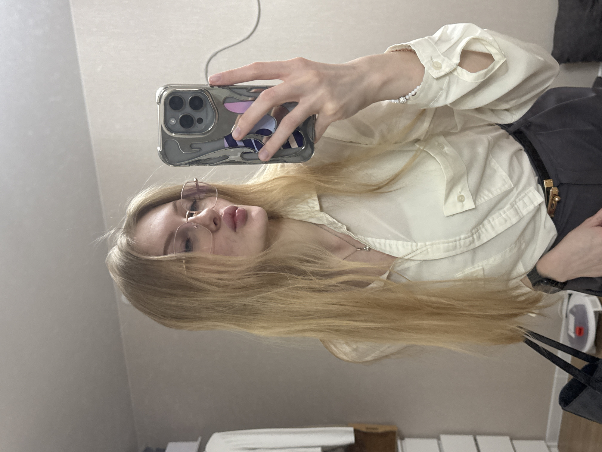 A blonde woman is taking a selfie in front of a mirror. She is wearing glasses, a white collared shirt, and a black belt. She is holding a phone with a distinctive phone case. The background is a light-colored wall, and portions of furniture are visible below, including what appears to be a set of drawers or a radiator. Due to lack of further context, there is no information regarding the location of the city of Perm, Russia, in this scene.