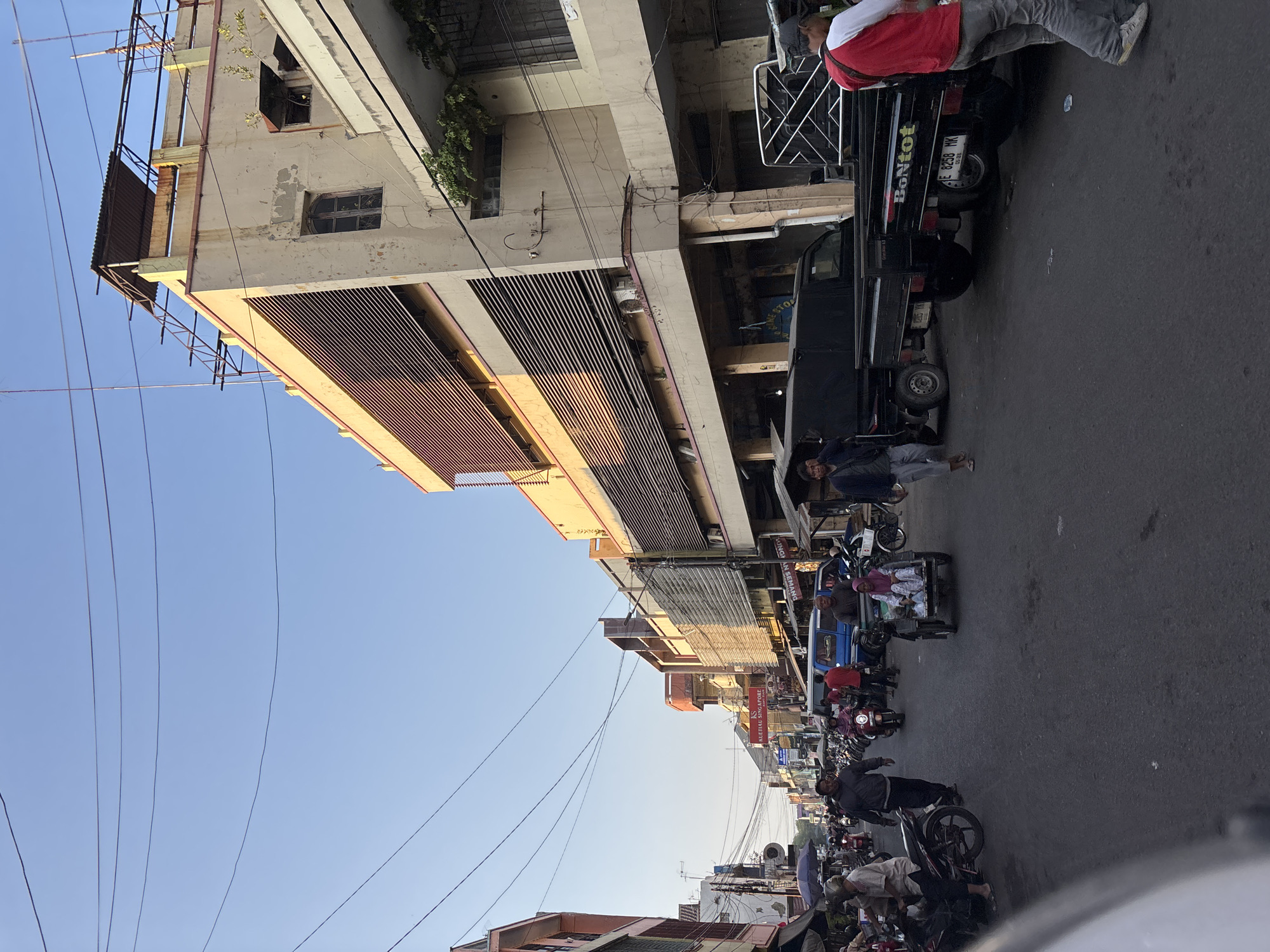 The image depicts a bustling street scene in Cirebon, Indonesia, likely within a commercial area. The time of day appears to be late afternoon, as indicated by the warm, angled sunlight illuminating the buildings on the right side of the street. The street is paved with asphalt and is filled with various forms of transportation.
On the right, a multi-story building with a light beige facade dominates the background. It features prominent horizontal louvers on its upper levels and some signs of wear and tear, with peeling paint and rusted metal elements on the roof. Adjacent to it, another building with a white lower section and an upper story with a dark metal-framed window is visible. Greenery is creeping up the facade of this building.
The street is crowded with motorcycles, some of which are parked along the sides, while others are in motion. A blue van is stopped in the middle of the street, and two dark-colored pickup trucks are also present. One of the pickup trucks has the word "BonTot" written on its tailgate.
Several people are present, walking or riding on the vehicles. On the left side of the street, market stalls or shops are lined up, with people browsing or tending to their businesses. A woman wearing a pink headscarf is seated in a three-wheeled vehicle, often used as a taxi in Indonesia. A man in a gray jacket walks towards the viewer, while another man in a red shirt and gray pants walks away.
The sky is a clear, pale blue, with multiple overhead electrical wires crisscrossing the frame, a common sight in urban areas of Indonesia. The overall impression is one of lively, everyday commerce and transit in a Southeast Asian city.