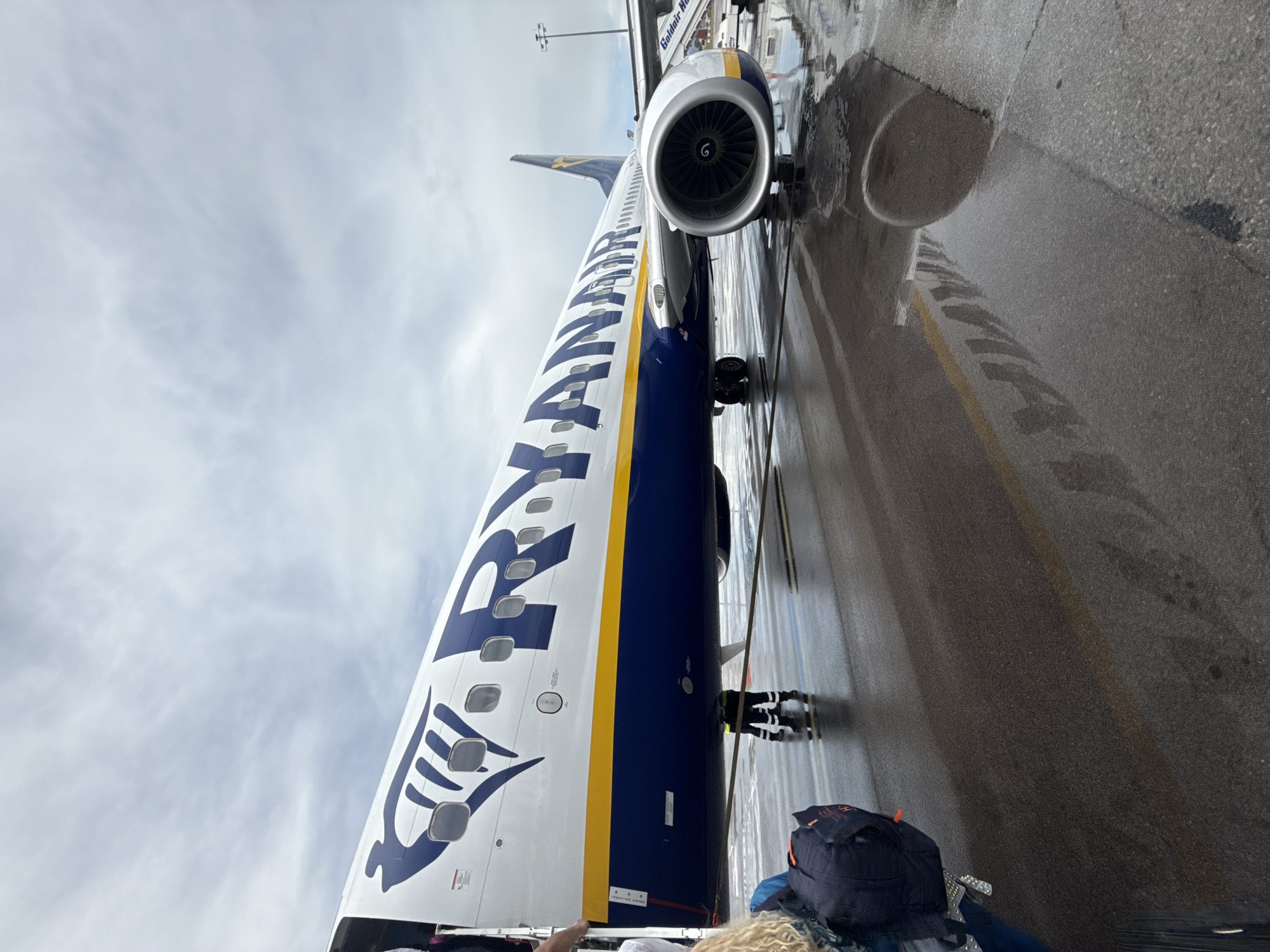 An eye-level shot captures a large blue and white Ryanair passenger airplane parked on a wet airport tarmac under an overcast sky in Sofia, Bulgaria. The aircraft's fuselage, emblazoned with the "RYANAIR" logo in large white letters and a distinctive yellow stripe, dominates the left side of the frame. The blue harp-like Ryanair logo is also visible towards the front of the plane.
The ground, made of asphalt, is noticeably wet with large puddles reflecting the plane's underside and the cloudy sky. One of the plane's engines, featuring exposed fan blades with a visible 'G' marking, is prominently seen in the middle-right. A ground crew member, wearing a high-visibility jacket and dark trousers, stands near the aircraft's landing gear, suggesting ongoing preparations for departure or arrival.
In the immediate foreground, a person's blonde curly hair and a dark blue backpack with orange accents are visible, indicating a passenger observing the scene. Part of an airport building or jet bridge structure with the partial text "Goldair Han" (likely "Goldair Handling") is visible in the background, further confirming the airport setting. The sky is a uniform, bright grey, typical of an overcast day.