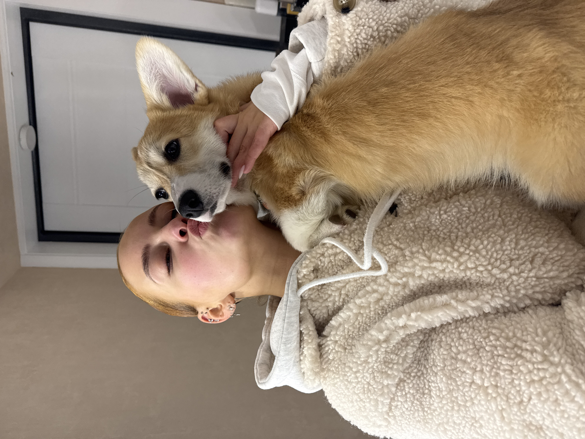 A woman is holding a corgi dog and kissing it on the nose. The woman has light brown hair and multiple piercings in her ear. She is wearing a white hoodie and a fuzzy white jacket. The corgi is mostly tan with white and black markings on its face. The background is a light brown wall and a white door with a black frame. The image was taken indoors, likely in a home, and is rotated to the left. It is not possible to confirm the location as Chaikovsky, Russia, without additional information.