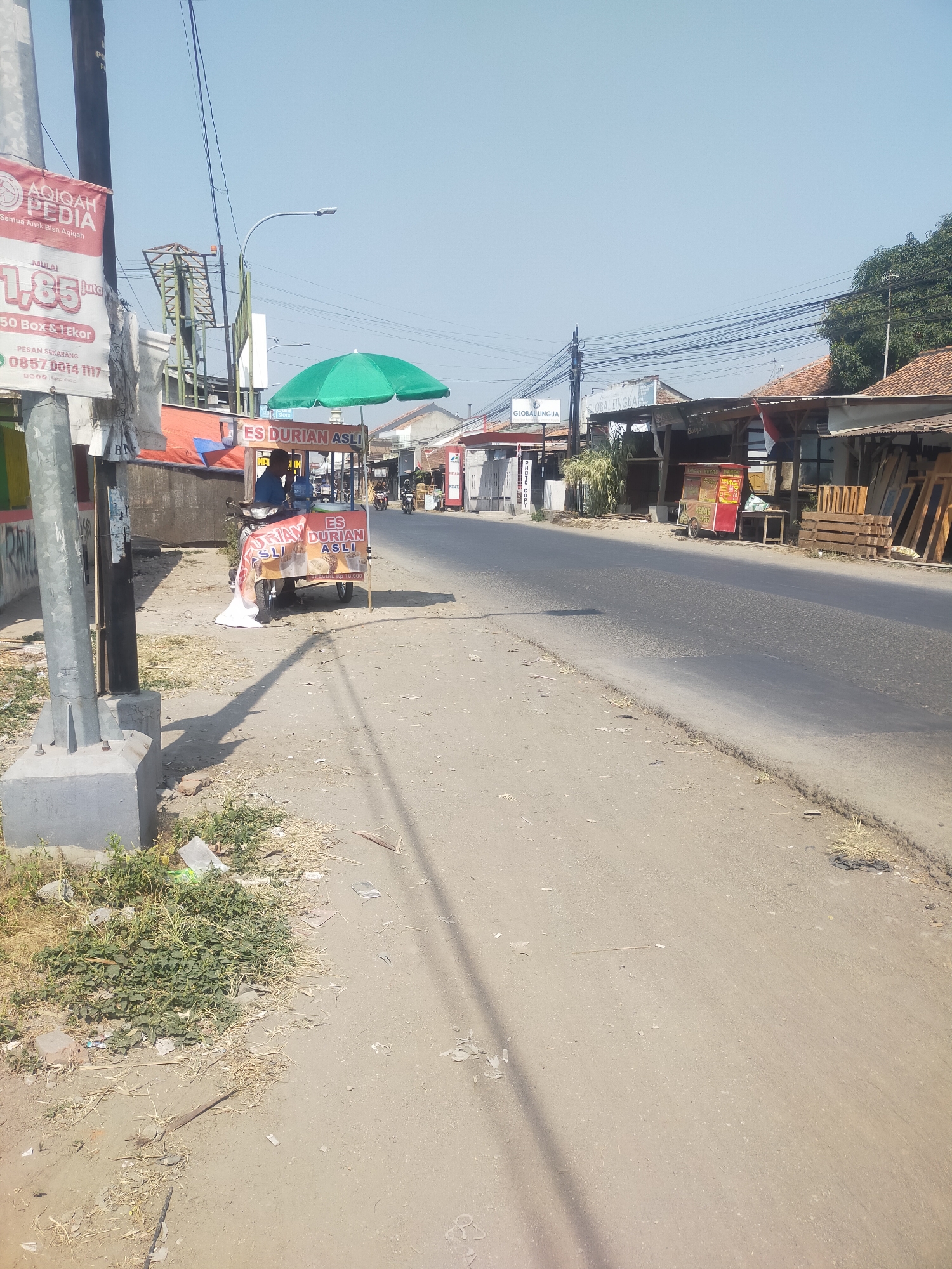 The image captures a street scene in Pegirikan, Indonesia, bathed in bright daylight under a clear blue sky. A prominent feature is a food cart selling "ES DURIAN ASLI" (Authentic Durian Ice) under a green umbrella. A person is visible behind the cart, presumably the vendor. The cart itself is adorned with images of durian and has text indicating the price.
To the left of the cart, a large red and white banner with the text "AQIQAH PEDIA" and "Semua Anak Bisa Aqiqah" (All Children Can Aqiqah) is attached to a utility pole. Another banner with "GLOBAL LINGUA" is visible further down the street. The street itself appears to be paved, with a dusty shoulder where the food cart is parked. Scattered debris, including plastic bags and dry vegetation, litters the roadside.
In the background, several buildings with tiled roofs line the street, suggesting a commercial or residential area. Electrical wires crisscross the sky, a common sight in urban Indonesian settings. The overall impression is of a typical day in a bustling Indonesian town, with street vendors and businesses operating alongside the road. The lighting suggests it is likely morning or early afternoon.