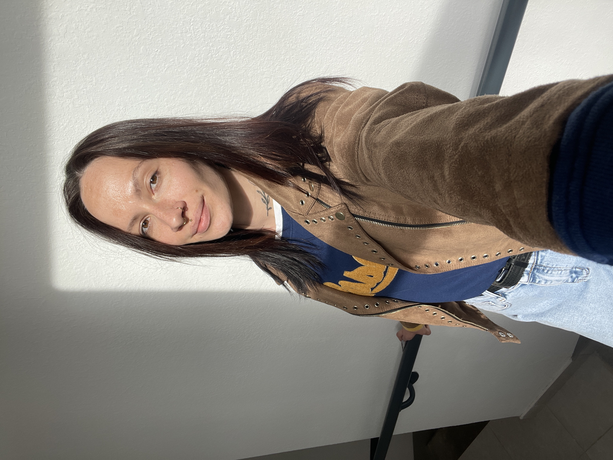 The image is a selfie of a young woman, likely taken indoors in Pervomaysky, Russia, under bright sunlight. The woman, with long dark brown hair, is looking down at the camera with a gentle, soft smile. Her face is illuminated by direct sunlight, which also casts prominent diagonal shadows on the light-colored wall behind her, suggesting a sunny day.
She is dressed in a brown suede-like jacket featuring multiple metal grommets or studs along the lapels and seams, worn over a navy blue long-sleeved shirt with a gold graphic design on the chest. She also wears light blue jeans and a black belt. Notable details include a septum piercing and a nostril piercing on her nose, and a small, delicate leaf or plant tattoo visible on her left collarbone/neck area. A yellow bracelet is visible on her right wrist.
The background consists of a plain white or light-colored textured wall, with a dark, possibly black, handrail visible on the right side of the frame. This suggests an indoor setting such as a hallway or staircase. No legible text is visible in the image. The overall impression is one of casual comfort in a well-lit environment.