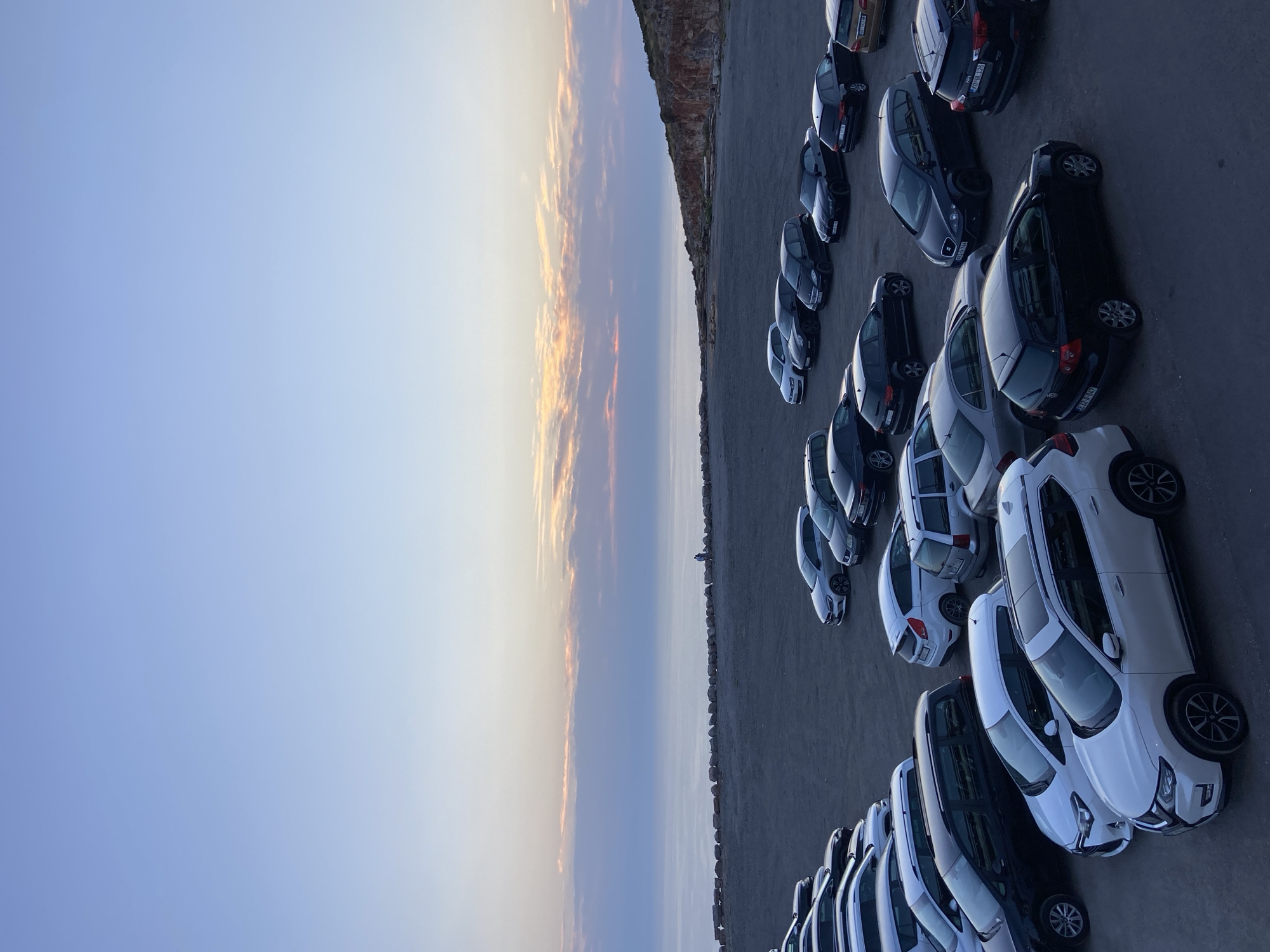 The image shows a parking lot filled with numerous cars, with a serene ocean and a dusky sky in the background. The parking lot is made of gravel and appears to be situated on a cliff or elevated area overlooking the sea. A stone wall separates the parking lot from the waterfront. In the distance, a single person is visible near the water's edge. The sky displays a gradient of blue and pastel hues, suggesting either sunrise or sunset. The cars are various makes and models, predominantly dark-colored, and parked in neat rows. The overall scene evokes a sense of calm and natural beauty, with the parking lot acting as a human-made element within a vast, natural landscape. The location is identified as Kovačko Polje, Montenegro.
