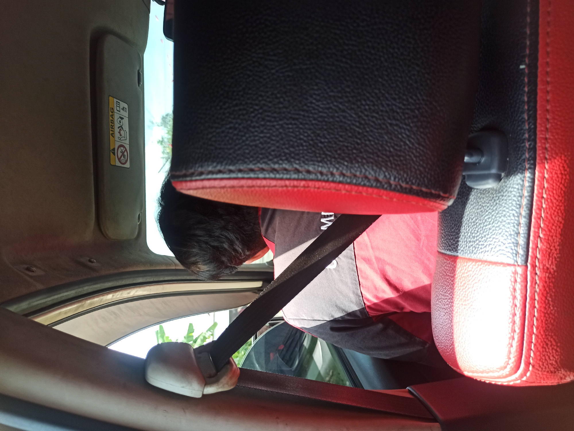 A person wearing a red and gray uniform with a seatbelt fastened is seen from behind inside a vehicle. The interior of the car features black and red leather seats and a sun visor with an "AIRBAG" warning label. Through the window, greenery is visible, suggesting an outdoor environment. The image was taken in the city of Medan, Indonesia, on a sunny day.