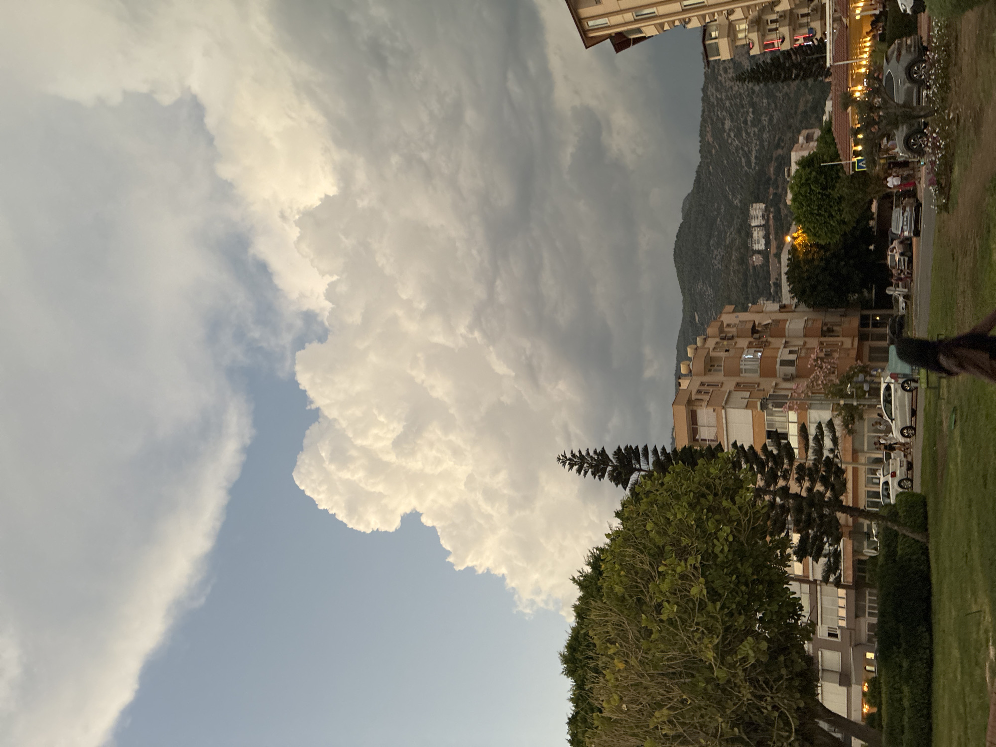 This image captures a street scene in Alanya, Turkey, likely during the late afternoon or early evening. The sky is dominated by dramatic, billowing white clouds that extend across much of the frame, contrasting with a lighter blue sky in the upper left. Below the clouds, a range of green, tree-covered hills forms the backdrop.
In the foreground and midground, several multi-story buildings line the street, with architectural styles suggesting a Mediterranean influence. These buildings have light-colored facades and numerous windows. One building on the right has a red-tiled roof and appears to be a commercial establishment, possibly a restaurant, with illuminated outdoor seating areas.
A variety of trees are visible, including a large, bushy tree with green foliage on the left and a tall, slender evergreen tree to its right. The street is populated with parked and moving vehicles, including several white cars and a dark gray SUV. A few people are present on the sidewalk and near the vehicles, though their interactions are not clearly discernible. One person in the lower right is partially visible, walking away from the camera.
The overall impression is that of an urban environment in a coastal town, with natural elements like the sky and hills playing a significant role in the composition. The lighting suggests a time of day when the sun is beginning to set, casting a warm glow on some parts of the scene.