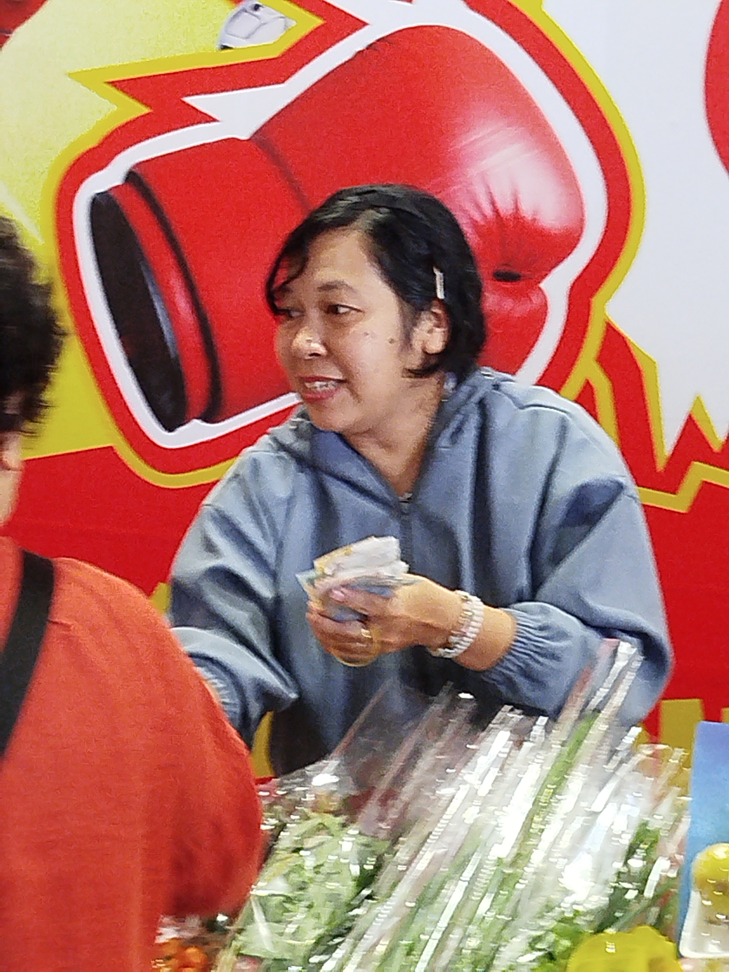 The image shows a woman in a blue hooded jacket standing behind a counter, holding a wad of cash in her hand. She is wearing a bracelet on her left wrist and a ring on her right ring finger. The background features a large, stylized graphic of two red boxing gloves with yellow and white accents. The woman appears to be smiling and looking to her right. In the foreground, there are various items on the counter, including what appear to be bundles of green vegetables and some yellow fruit. The scene suggests a commercial transaction is taking place, possibly at a market or shop. The image's context of Da Lat, Vietnam, implies this could be a local market or a roadside stall in the city.