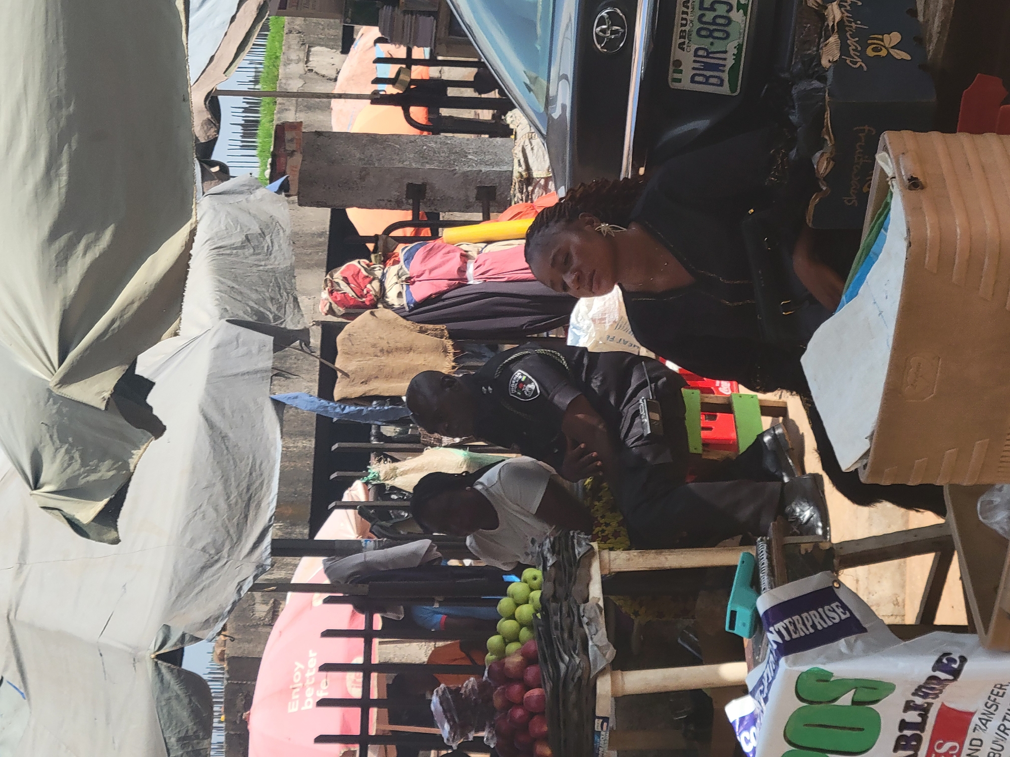 This image captures a vibrant street market scene in Abuja, Nigeria, during the daytime. The perspective is from within the market, looking out towards a road and other stalls.
In the foreground and midground, market stalls are covered by large, light-colored tarpaulins or canvas sheets, providing shade. On the right side, a black car, identified as a Toyota by its emblem, is parked. Its license plate clearly reads "ABUJA," "CENTRE OF UNITY," and "BWR-865," confirming the location.
Several individuals are visible. Closest to the car, on the right, a woman with braided hair and a distinctive star-shaped earring is wearing a dark top. She appears to be sitting or leaning, looking downwards with a neutral expression. Behind her and slightly to the left, a man in a dark uniform with insignia, possibly a police officer, is also seated or bent over, observing something. Further left and behind the uniformed man, another person in a white top is partially visible near a stall laden with green and red apples.
The market environment is characterized by various goods and makeshift structures. Below the car, a blue sign with a bee logo and the word "fruitways" is attached to a crate. Other containers include cardboard boxes and a white plastic bag with "ENTERPRISE" and "S" prominently displayed, along with fragmented text like "ABLEHORE," "ND WANSFER," and "MEAT FE." A partial view of a red surface, possibly an umbrella or canopy, carries the text "Enjoy better fe." The overall impression is that of a bustling local market under sunny skies.